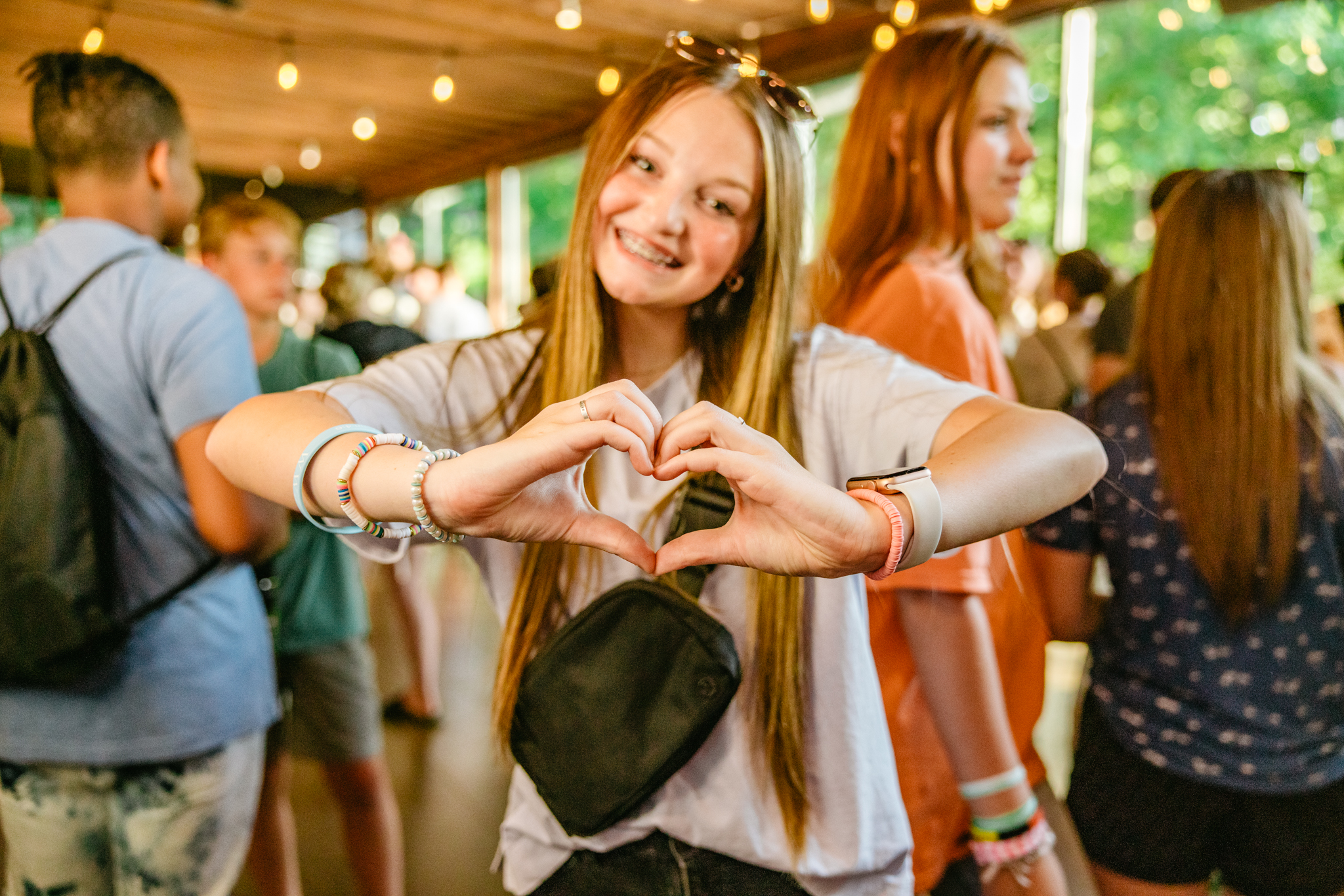 Teen girl smiling with braces forms a heart shape with her hands at a crowded indoor event.