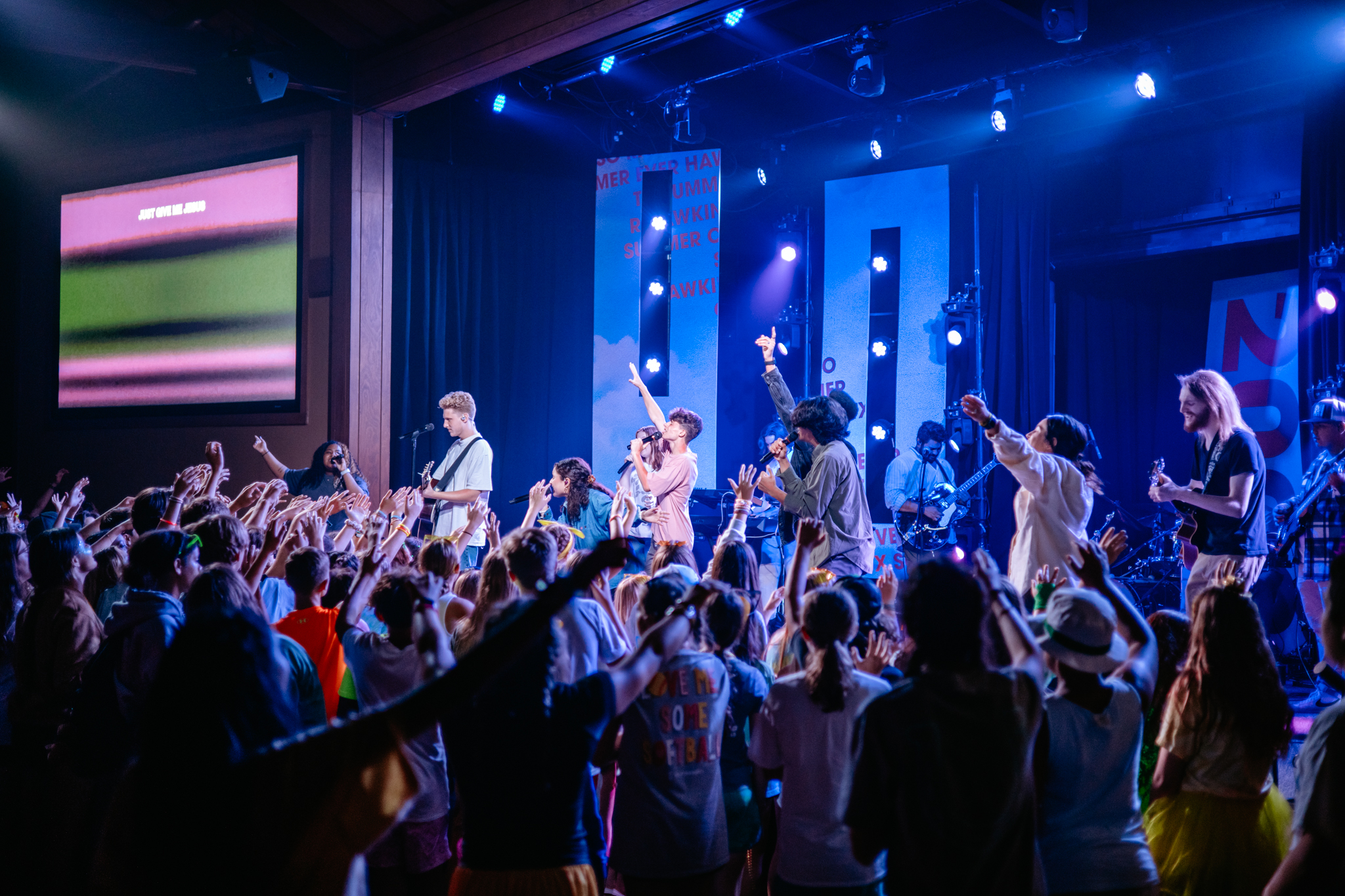 Crowd of young people with raised hands enjoying a live band performing on a stage with colorful lights.
