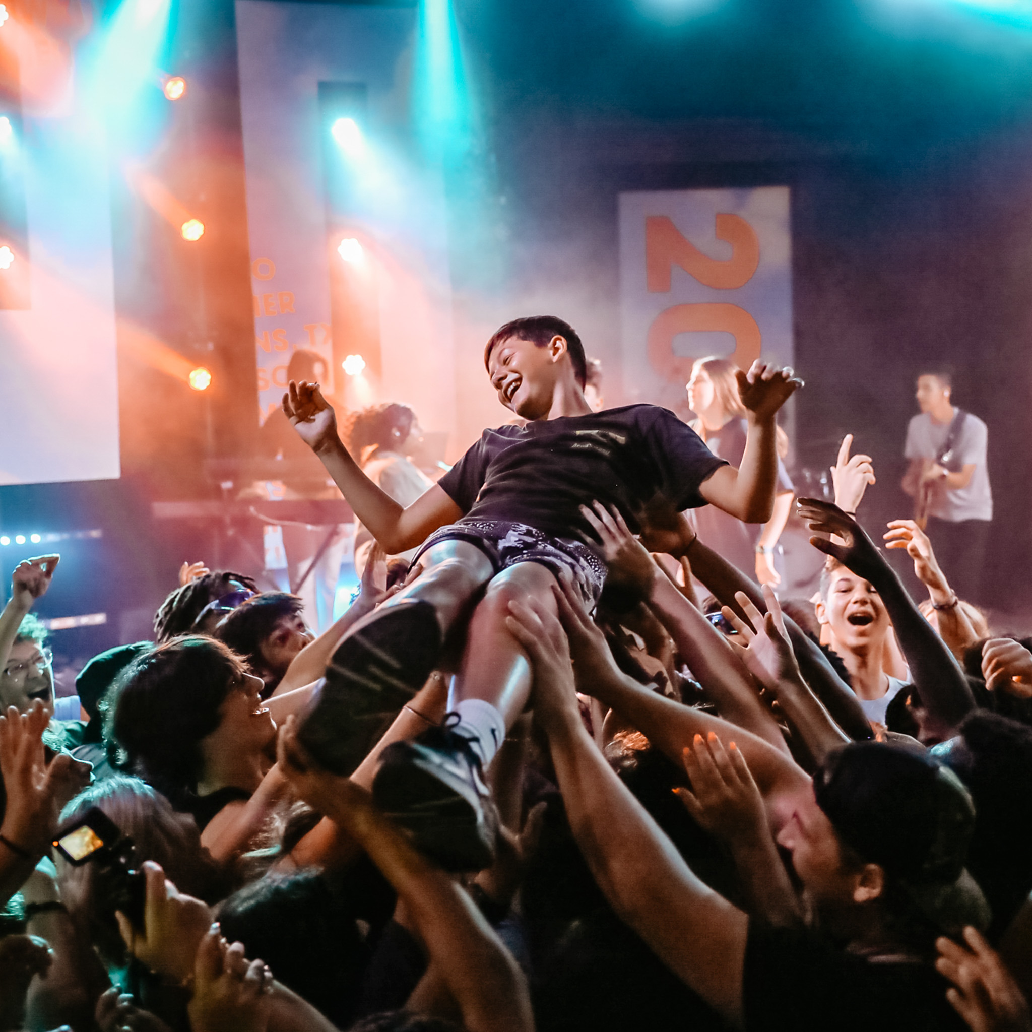 A young boy crowd surfing over a cheering audience at a lively indoor concert with colorful stage lighting.
