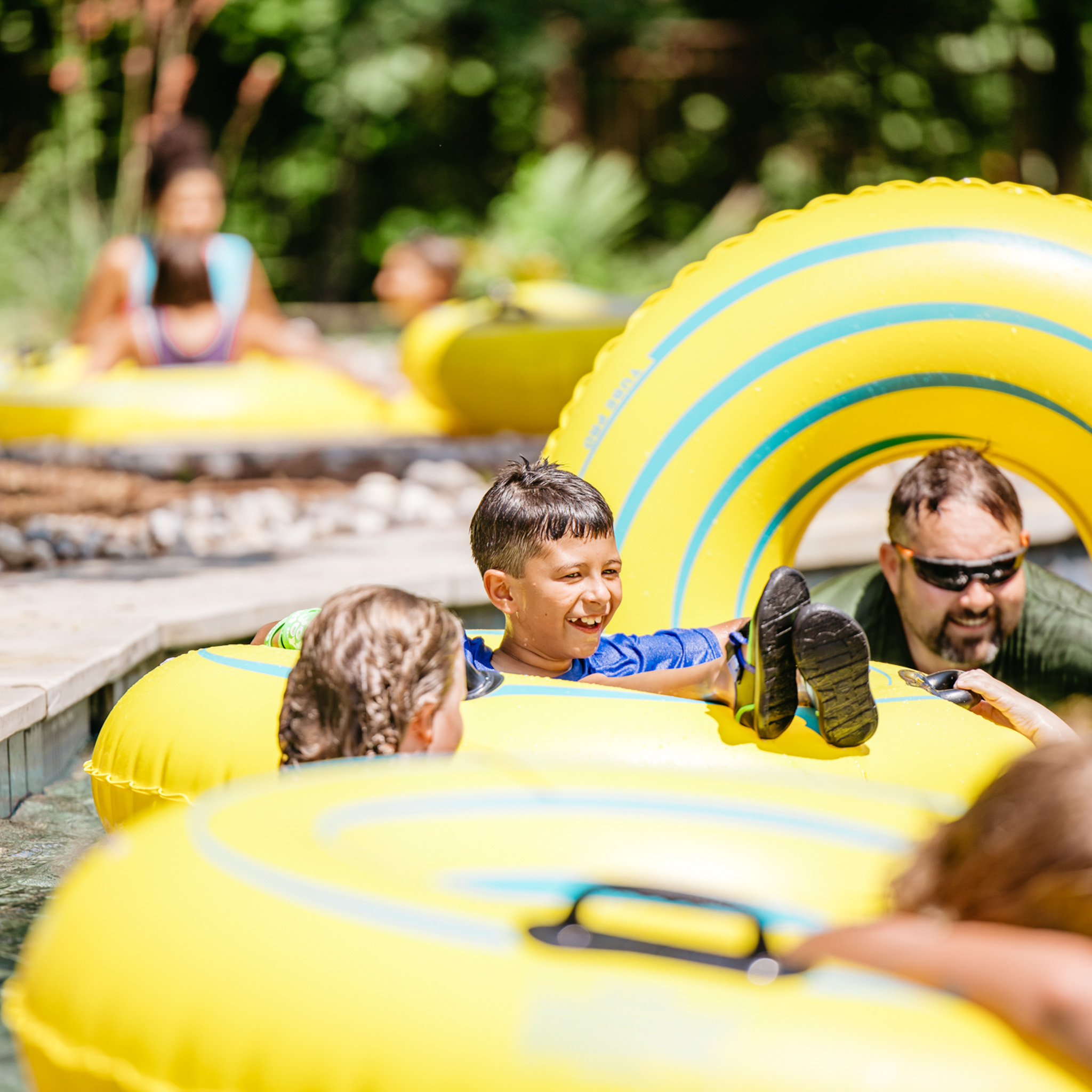 Smiling boy wearing a blue shirt relaxing in a yellow inflatable tube in a pool with other people.