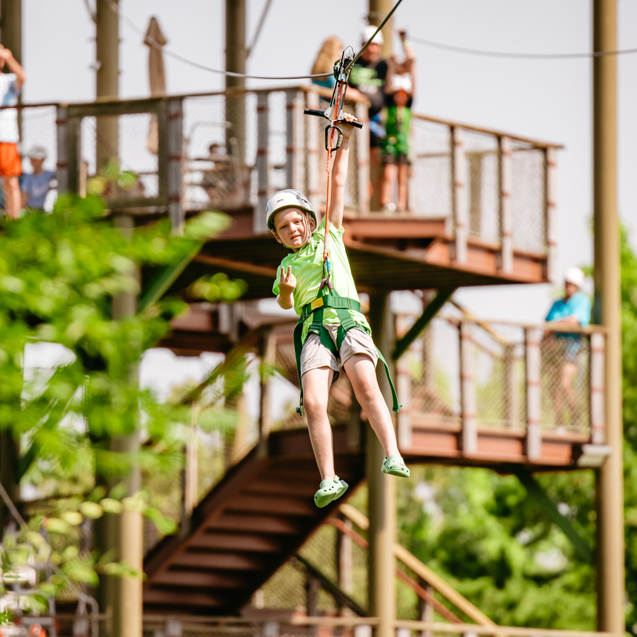 Child wearing a helmet and harness rides a zip line past a wooden platform with people watching.