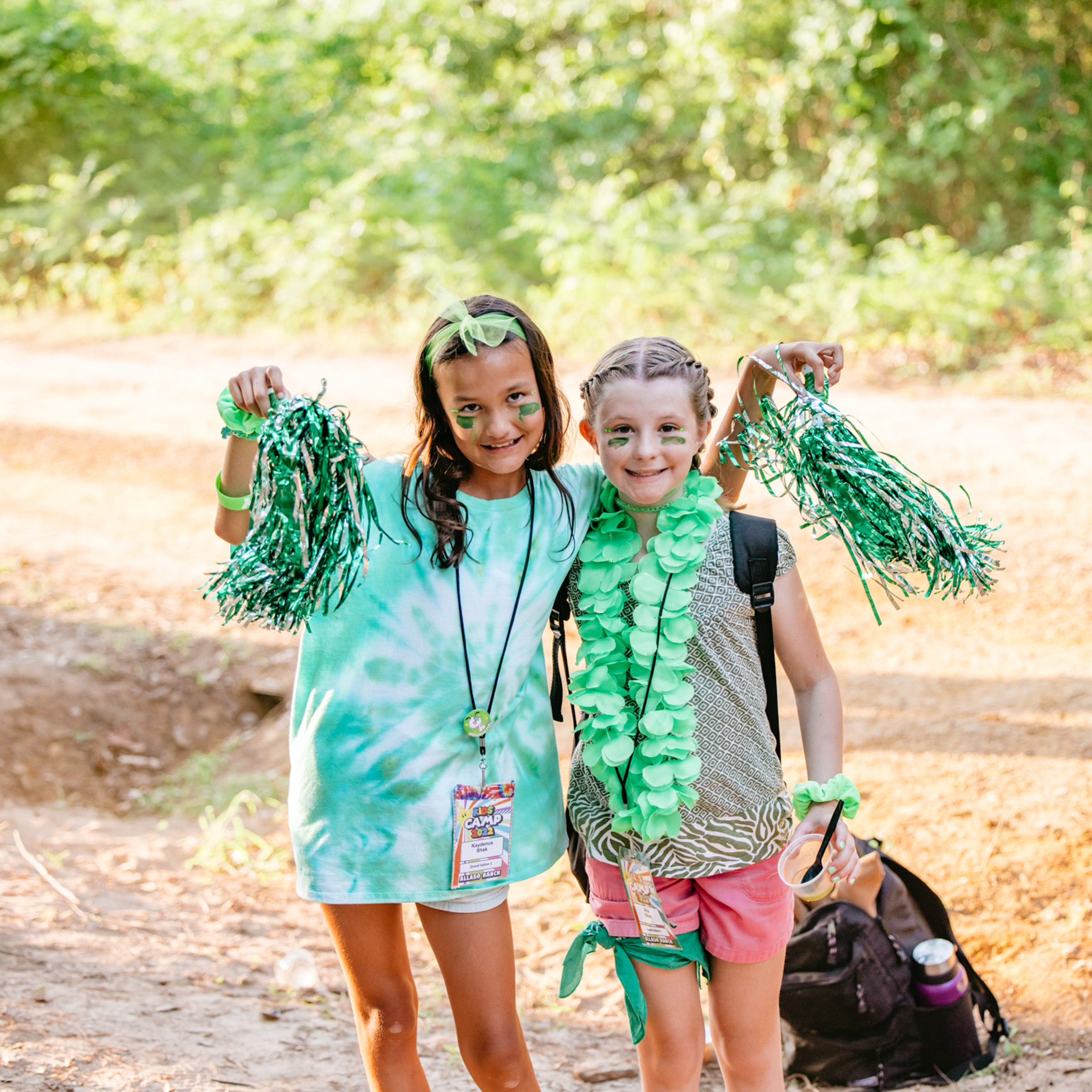 Two smiling girls outdoors wearing green accessories and holding green pom-poms, with nature in the background.