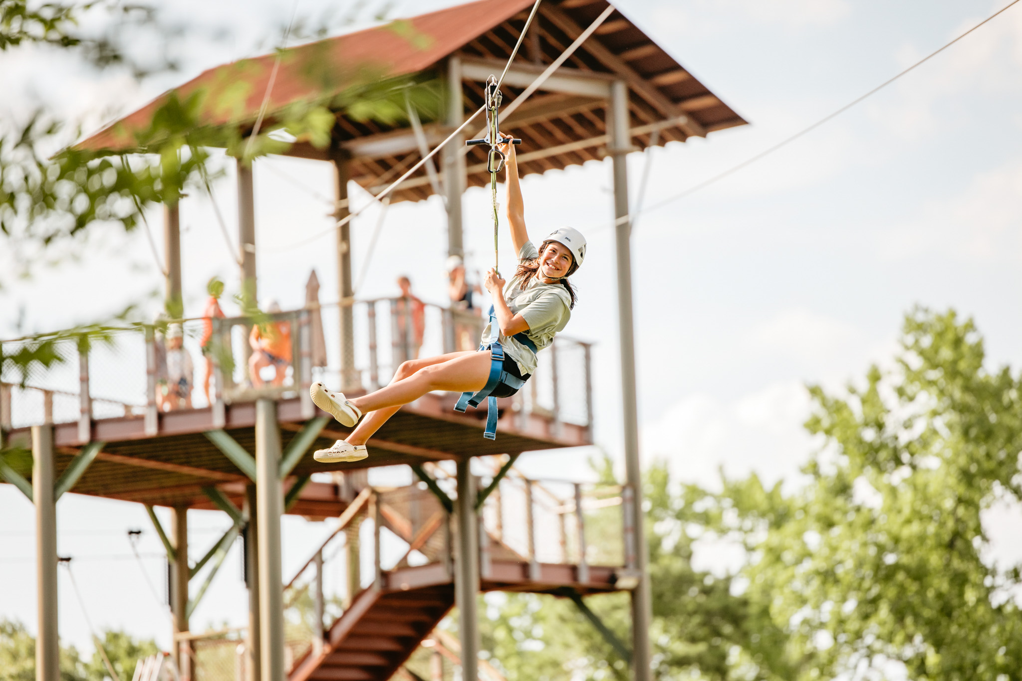 Smiling woman wearing helmet and harness riding a zip line with wooden platform and trees in the background.