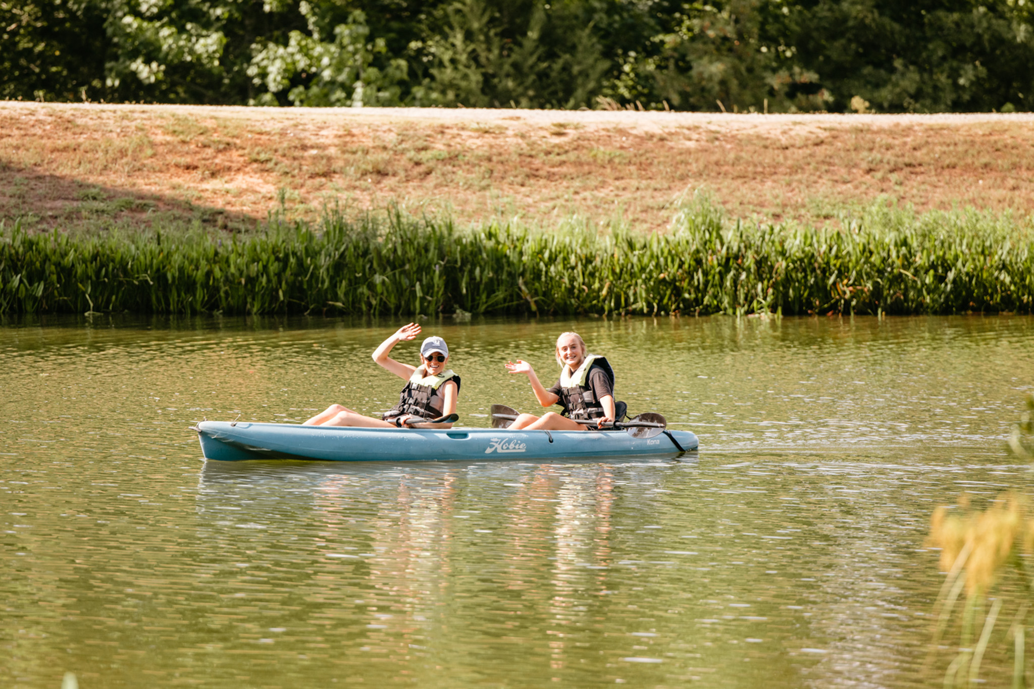 Two people wearing life jackets waving while sitting in a blue kayak on calm water surrounded by greenery.