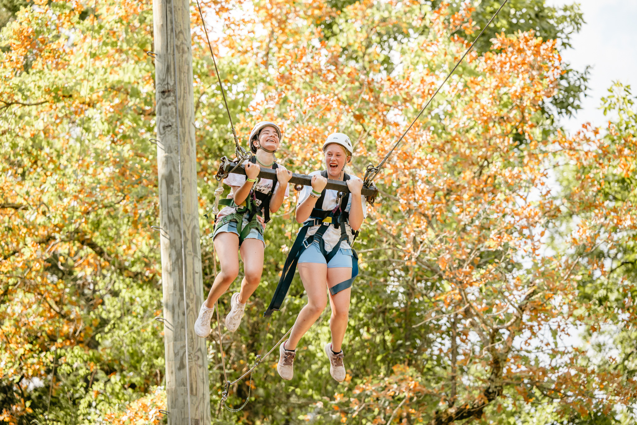Two girls wearing helmets and harnesses laughing while suspended on a large swing against autumn foliage.