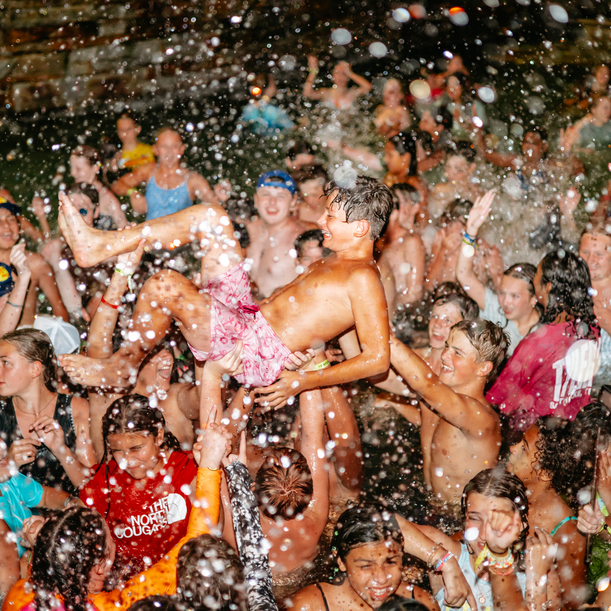 A group of children splashing water and lifting a boy in pink swim trunks above the crowd, all enjoying a lively water party.