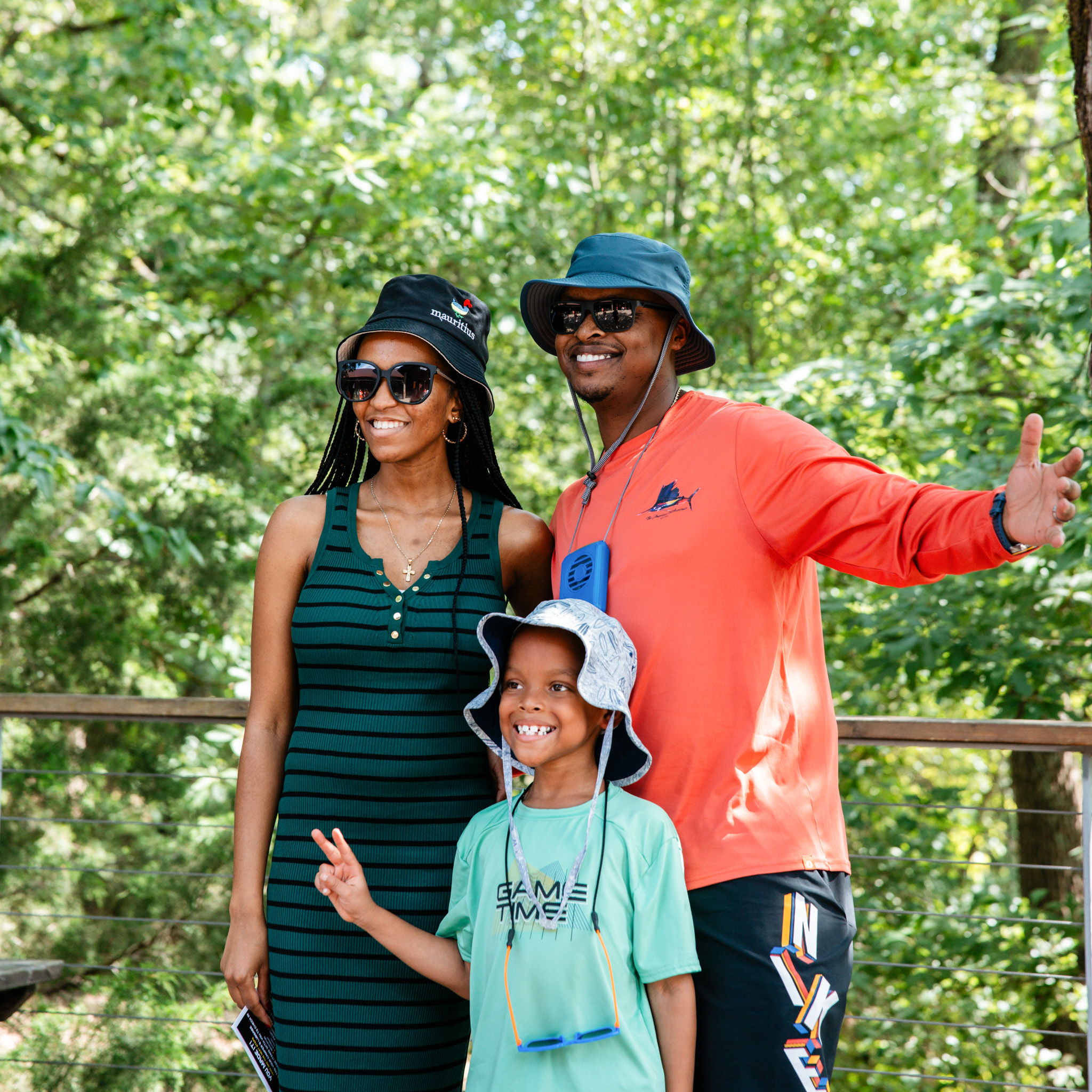 Smiling family of parents and one child wearing sun hats outdoors surrounded by trees.