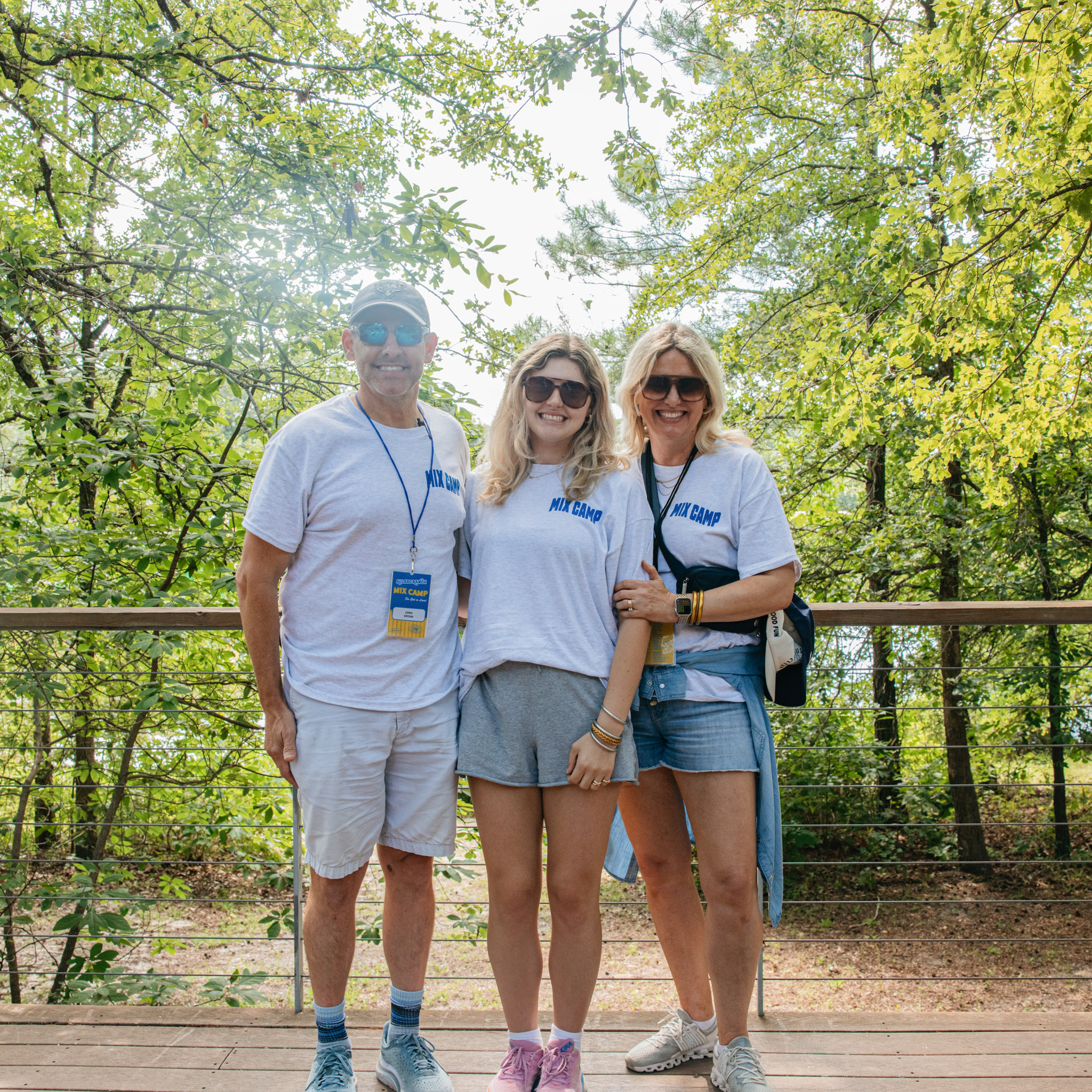 Teenage girl and her parents wearing matching white MIX CAMP t-shirts and sunglasses standing on a wooden deck surrounded by green trees.