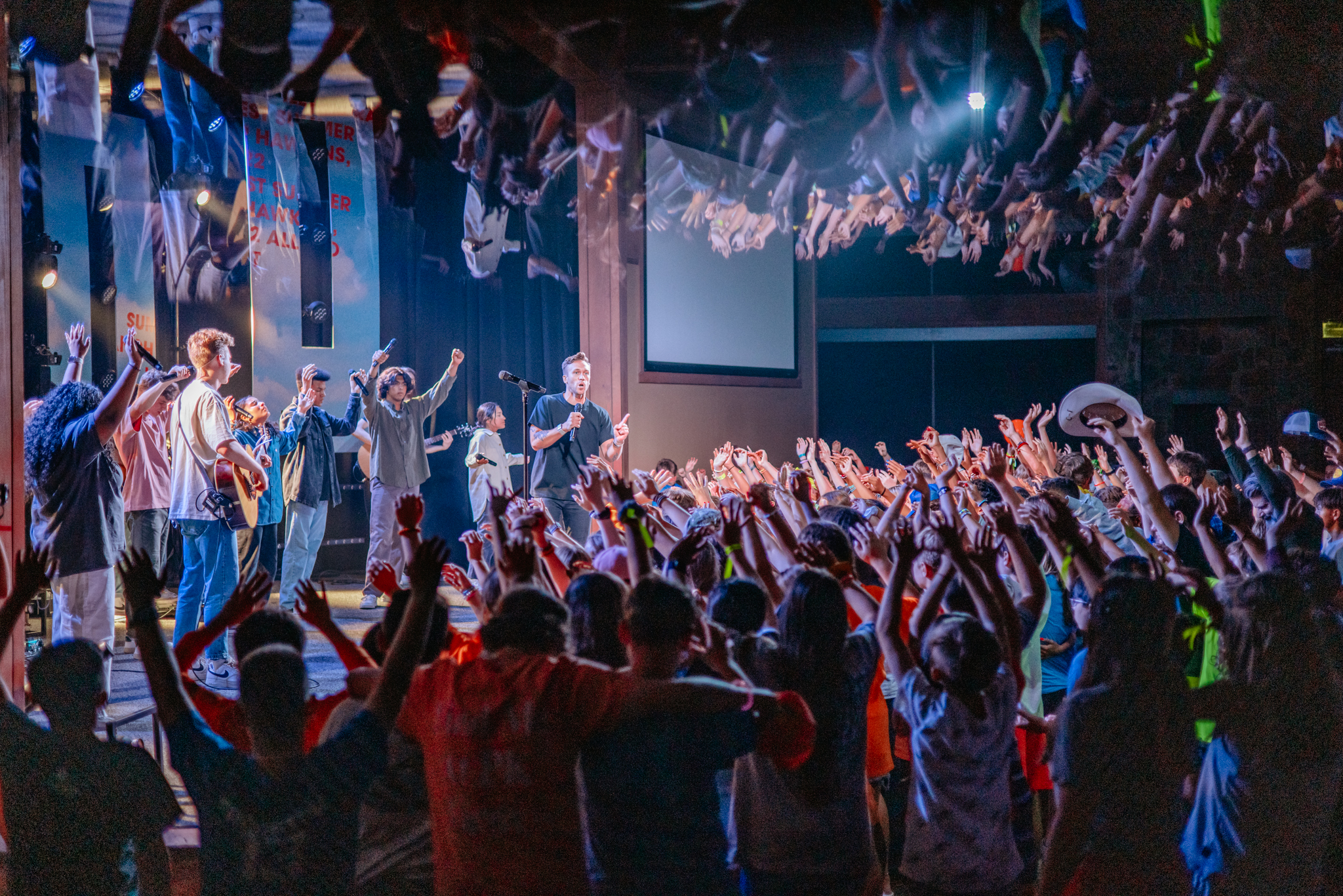 Crowd with raised hands participating in an indoor event with a speaker and musicians on stage.