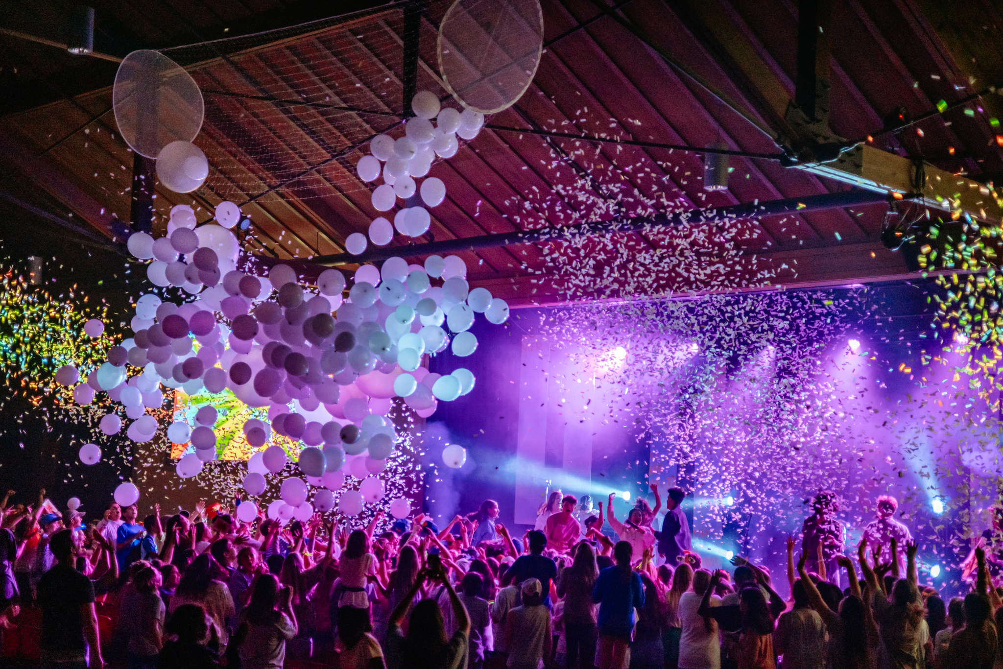 Crowd celebrating indoors with white balloons and colorful confetti under purple stage lights.