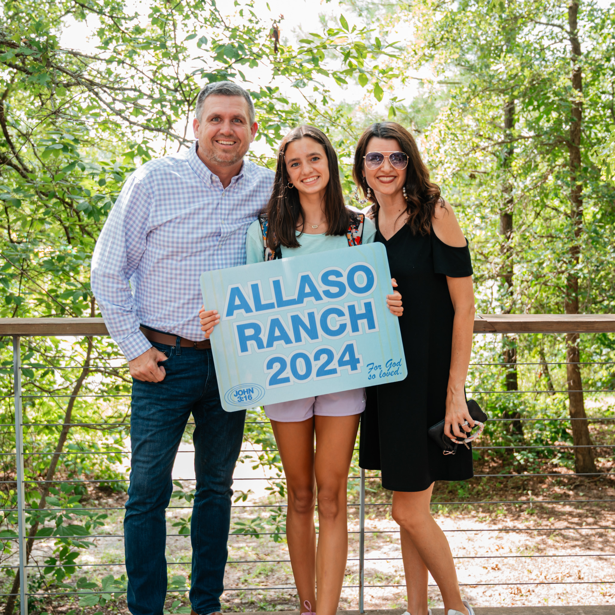 Smiling family of three outdoors holding a sign that reads 'Allaso Ranch 2024' with trees in the background.