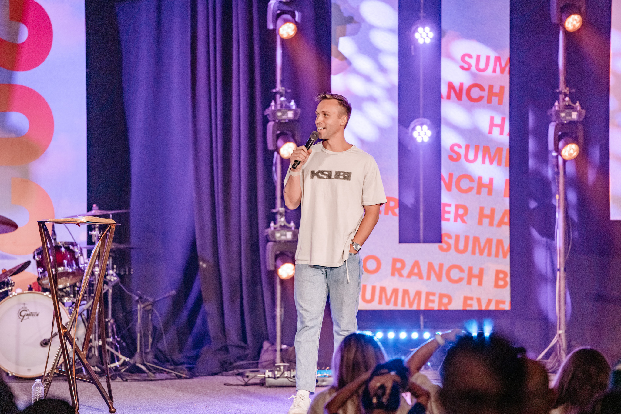 A young man holding a microphone and speaking on stage in front of an audience, with musical instruments and stage lights in the background.