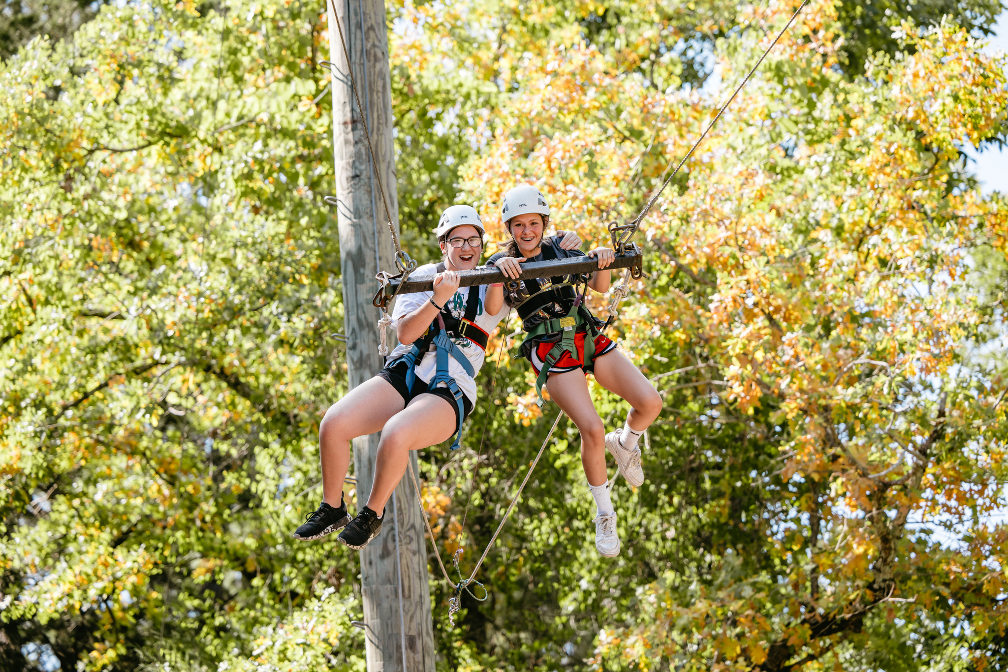 Two girls wearing helmets and harnesses smiling while suspended on a high ropes course with green and yellow foliage in the background.