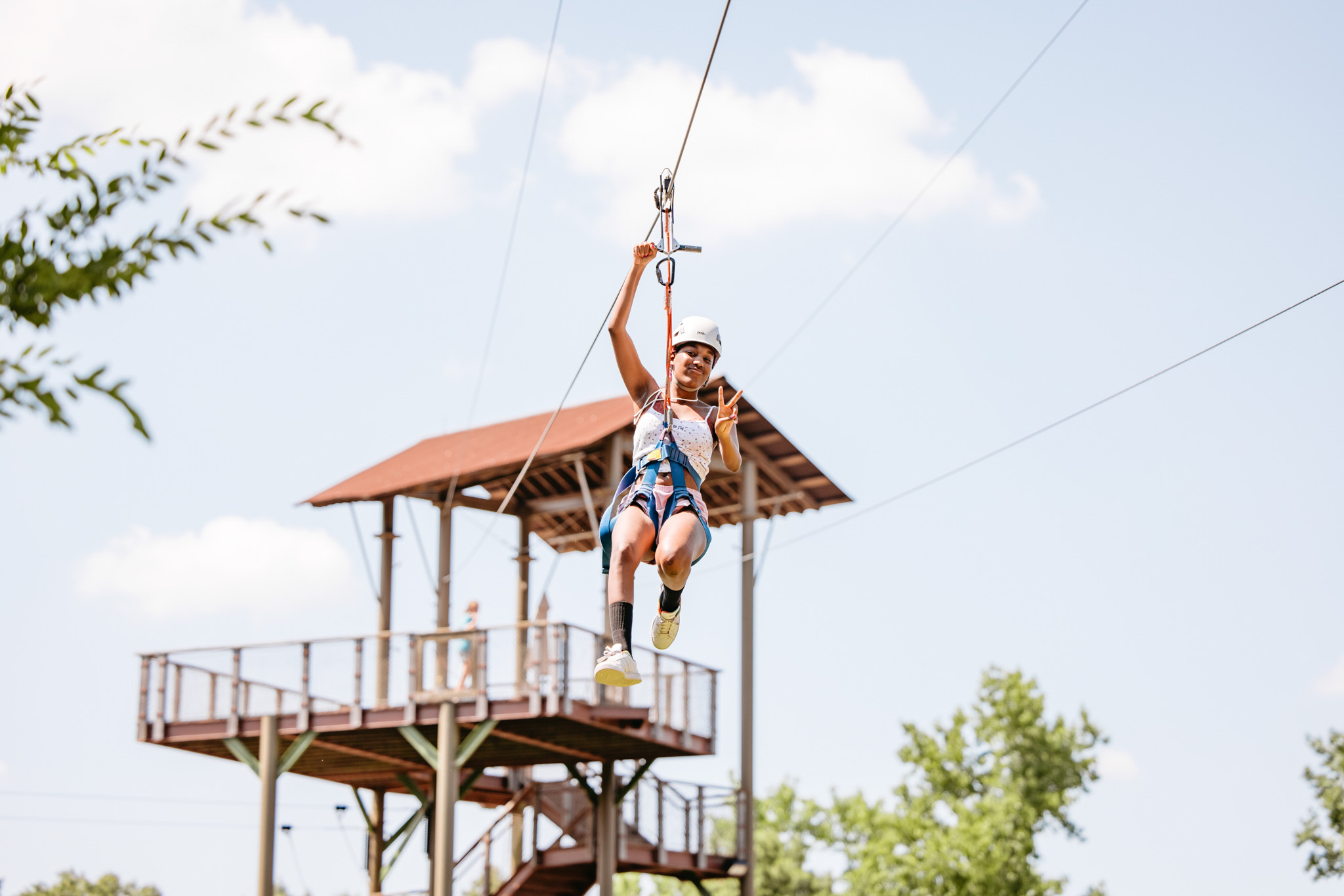 Young woman wearing a safety helmet and harness making a peace sign while riding a zipline with a wooden platform in the background.