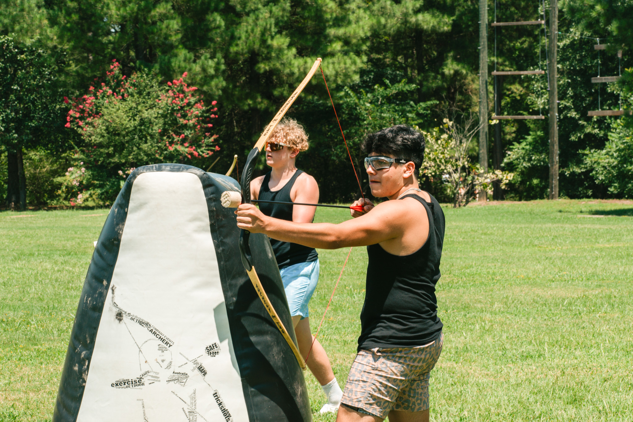 Two young men practicing archery outdoors on grass, aiming at a large padded target.