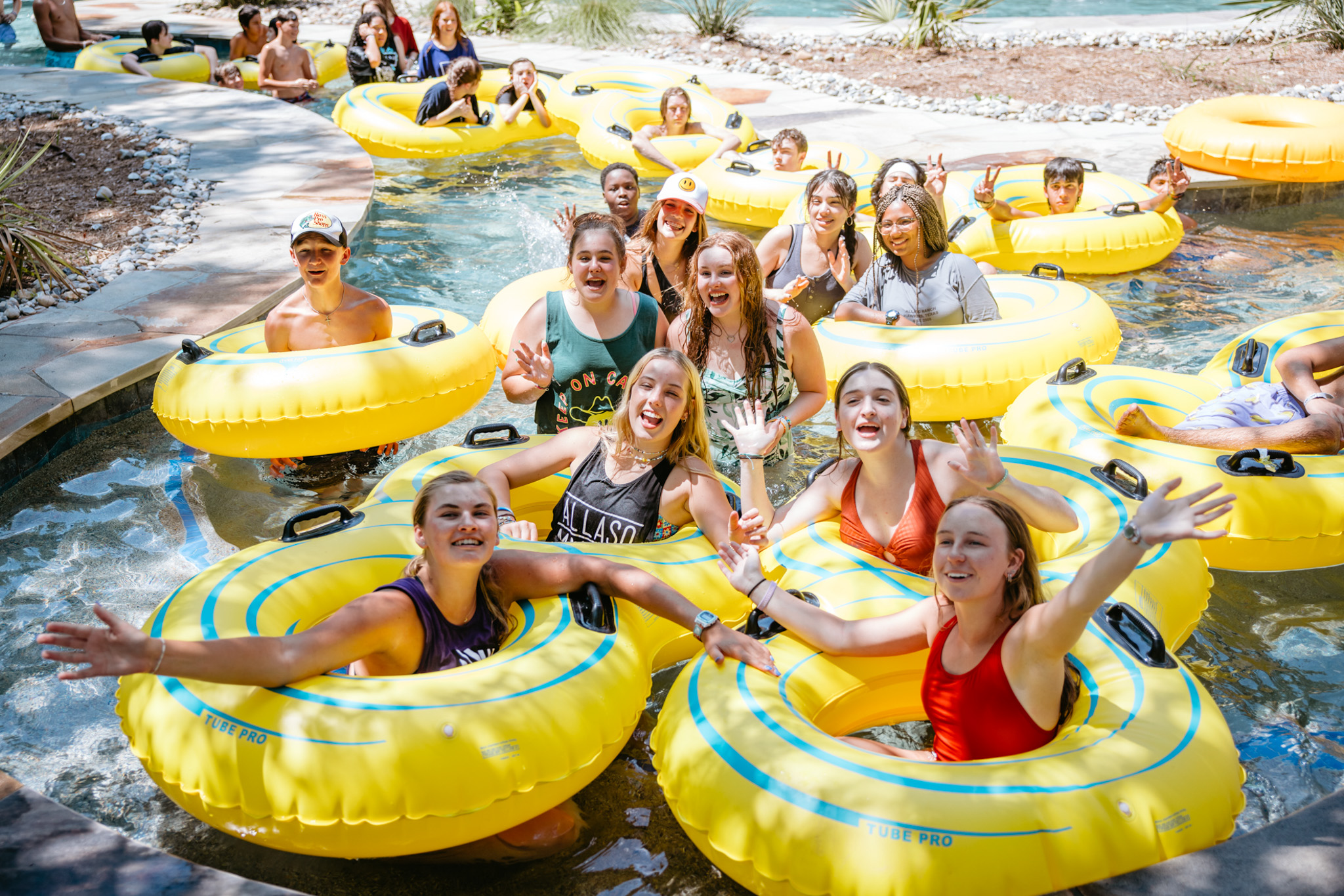 Group of smiling young people enjoying a lazy river with yellow inflatable tubes on a sunny day.