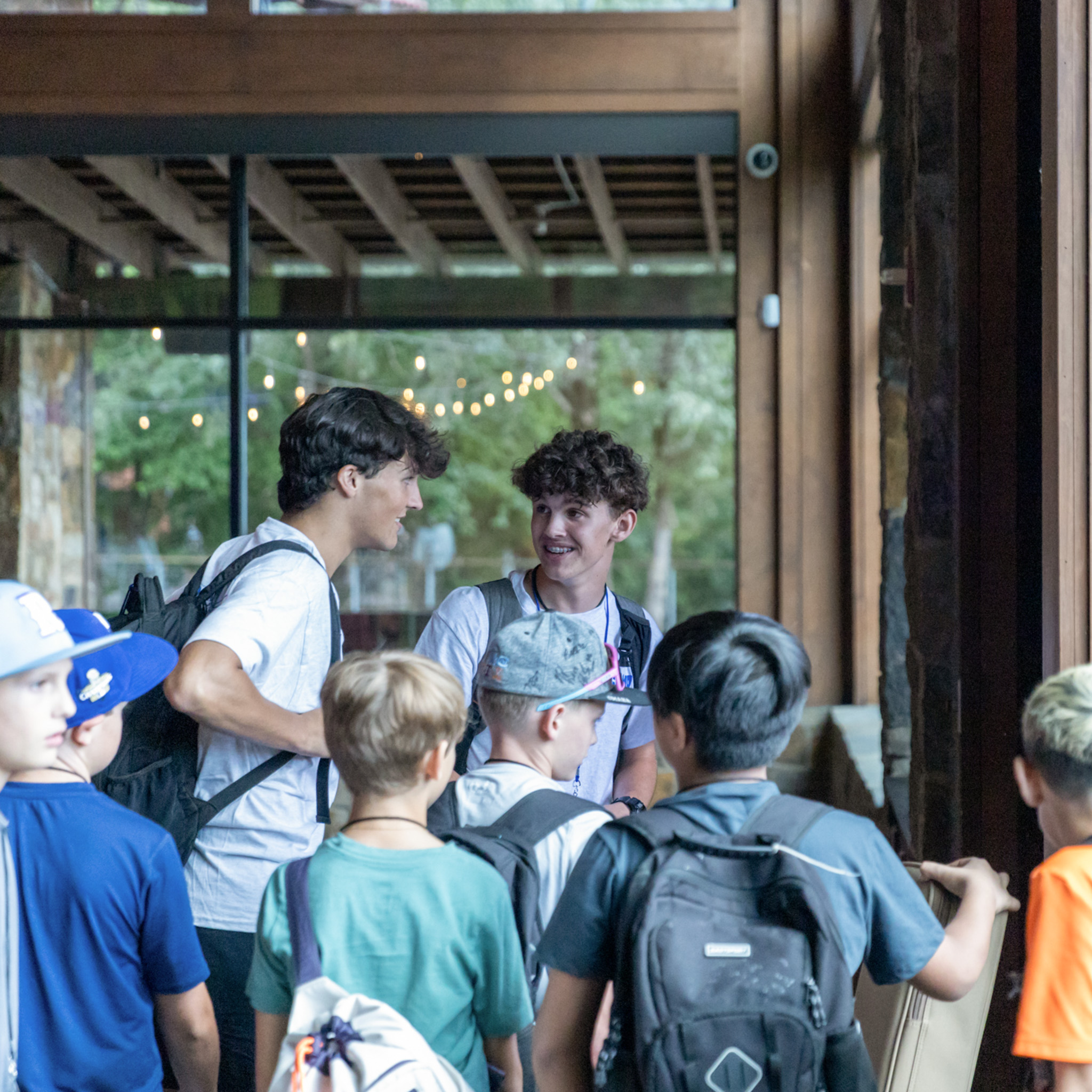 Two teenage boys with backpacks smiling and talking to a group of younger boys near a wooden doorway.