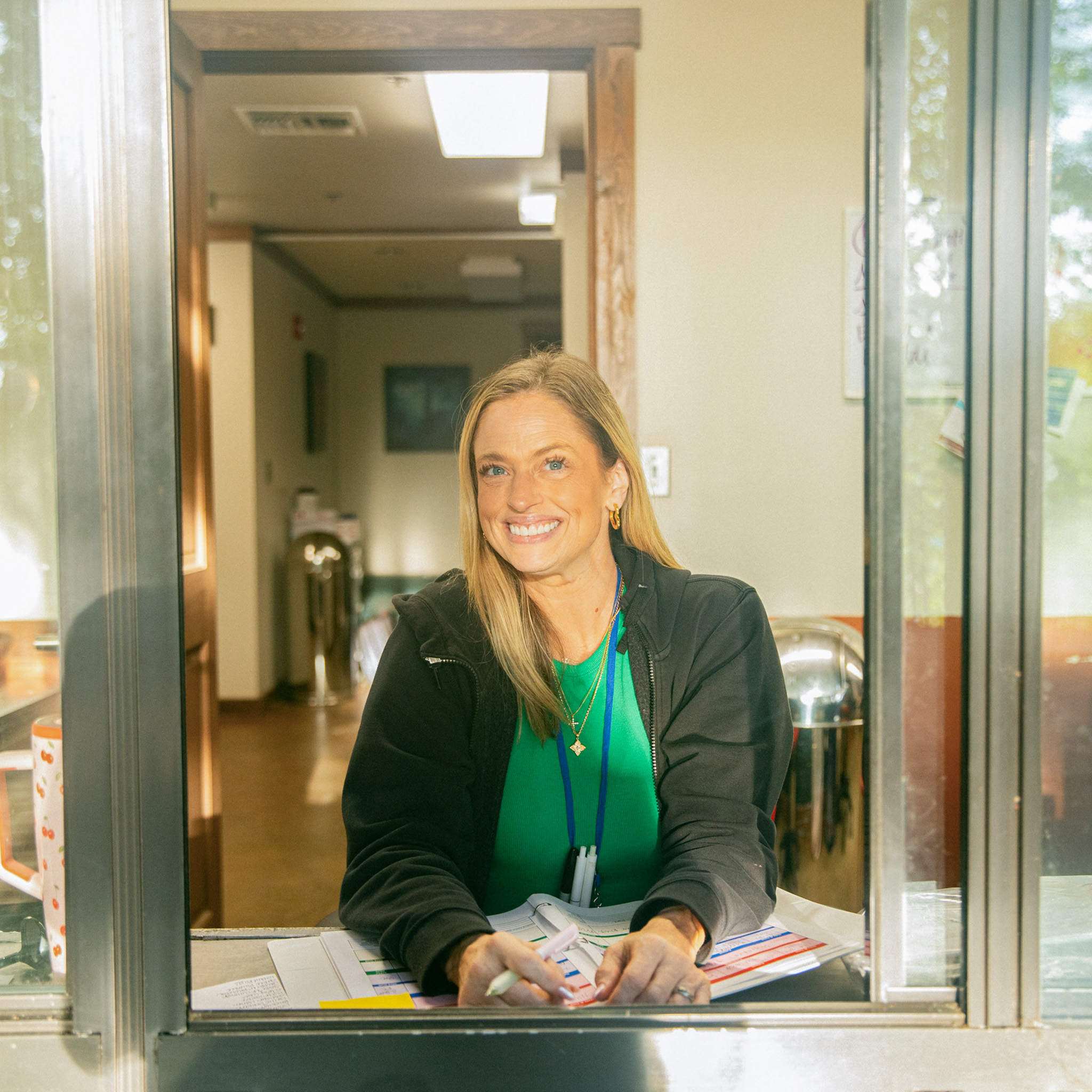 Smiling woman with blonde hair wearing a black jacket and green shirt, sitting at a counter with papers and pens.