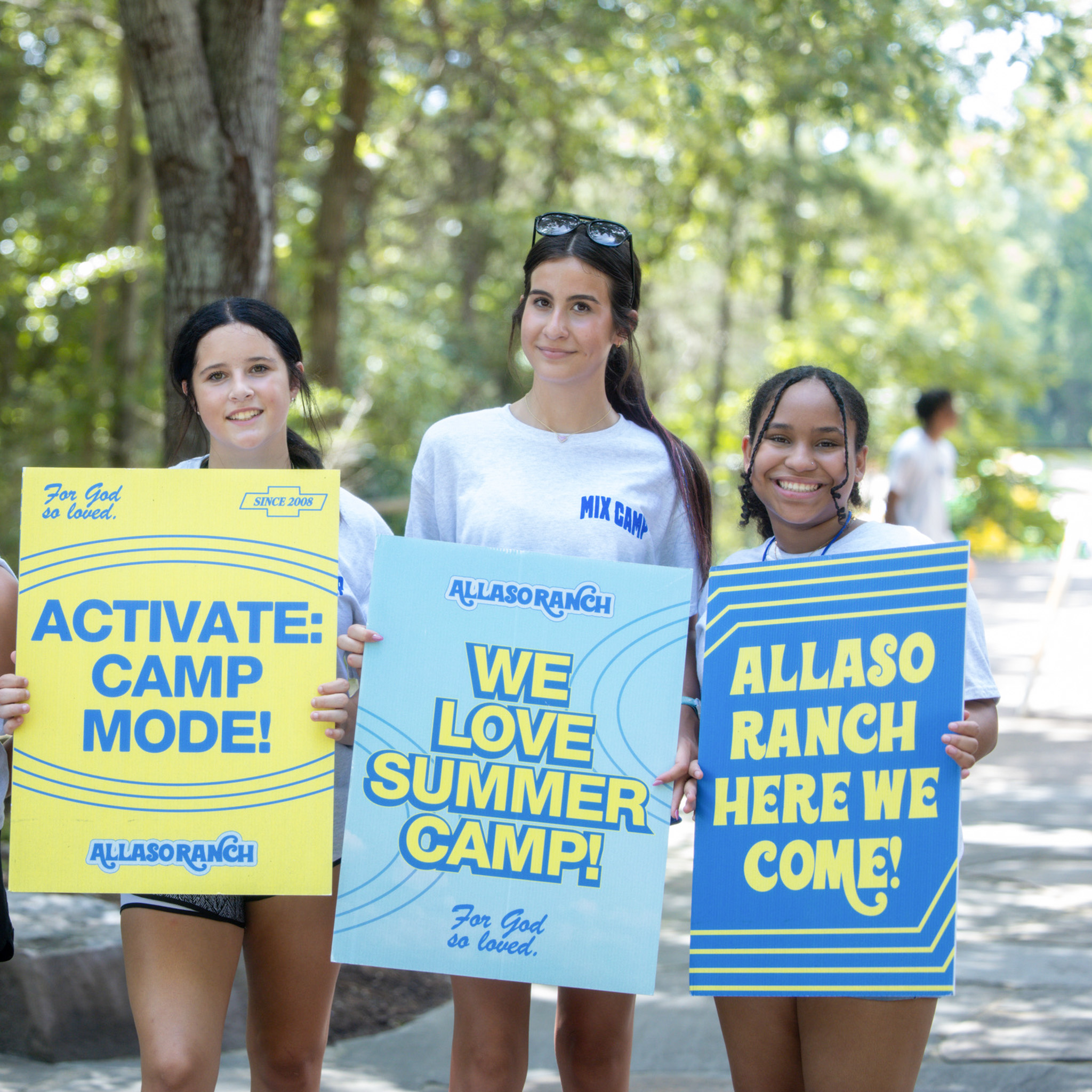 Three smiling young women holding colorful signs with messages supporting Allaso Ranch summer camp outdoors.