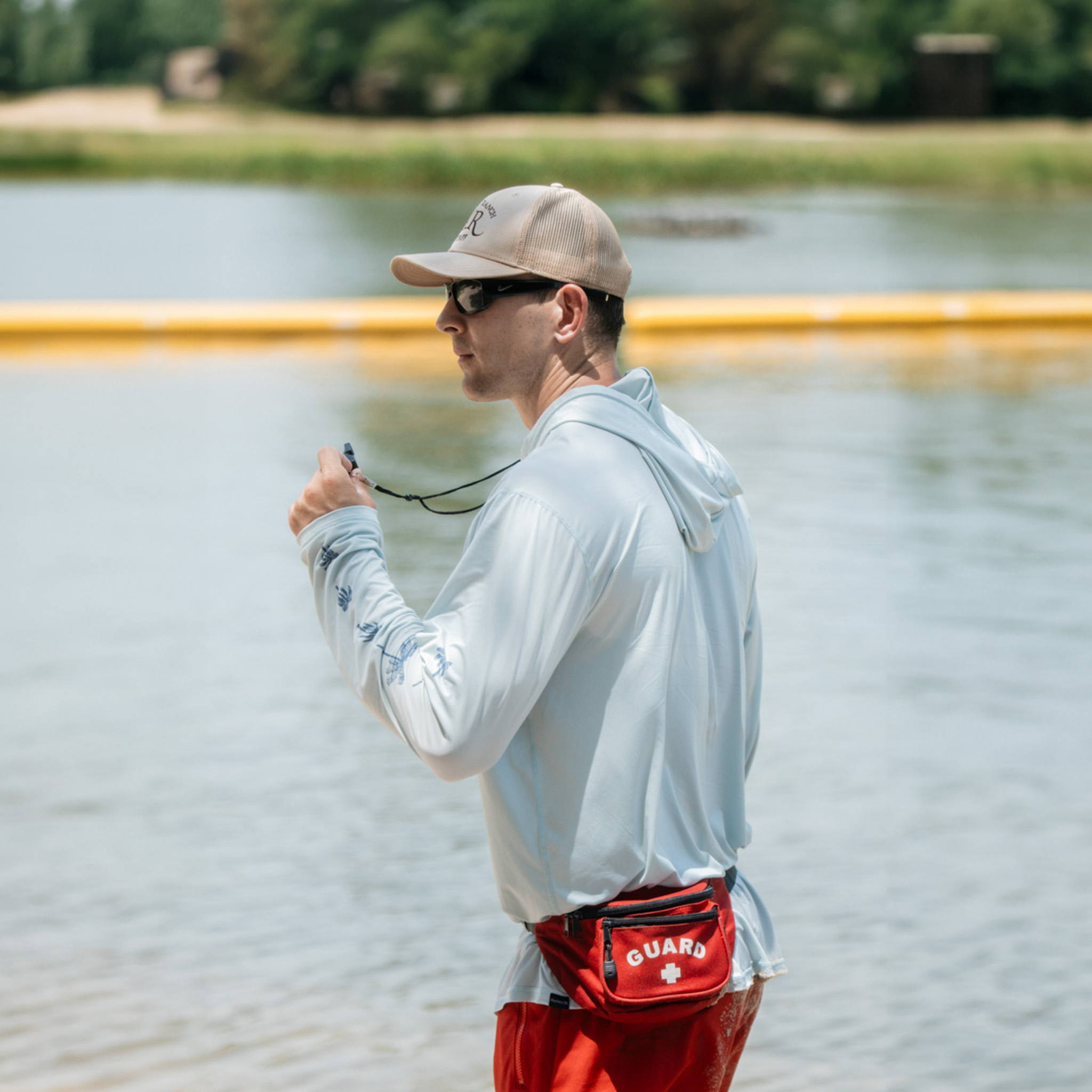 Lifeguard in sunglasses and cap standing by a body of water holding a whistle.