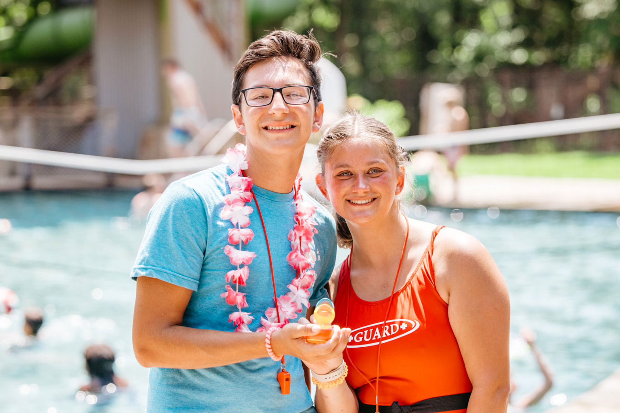 Smiling lifeguards standing outdoors near a pool, one wearing a red lifeguard swimsuit and the other a blue shirt with a floral lei.