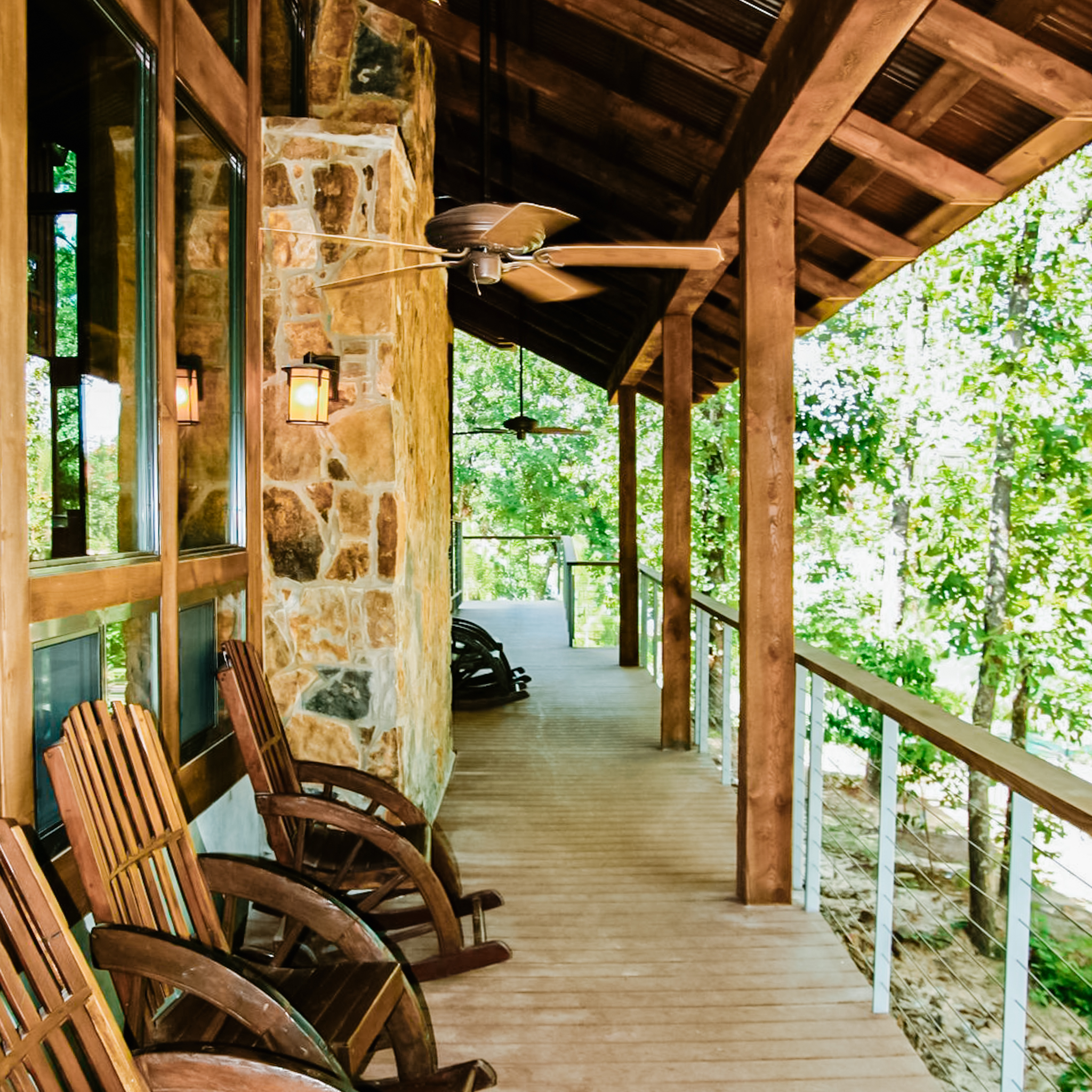 Wooden porch with rocking chairs, ceiling fans, and stone wall surrounded by trees.