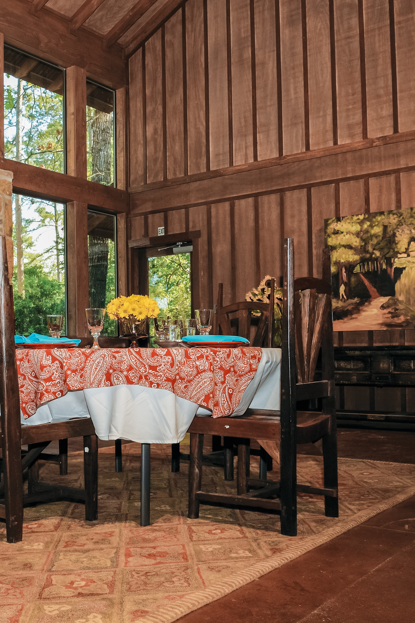Dining table with orange paisley tablecloth and turquoise napkins in a wooden cabin with large windows showing green trees outside.