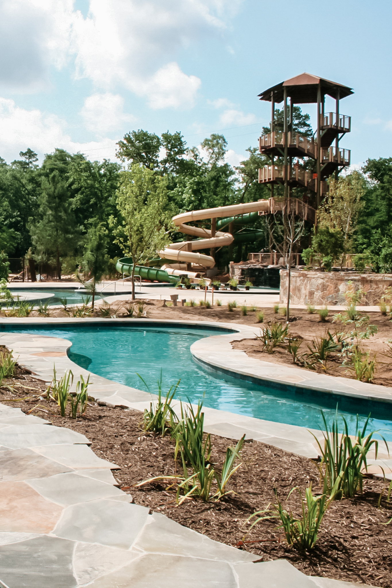 Outdoor water park with winding pool, surrounded by stone walkway and plants, and tall brown water slides in the background.