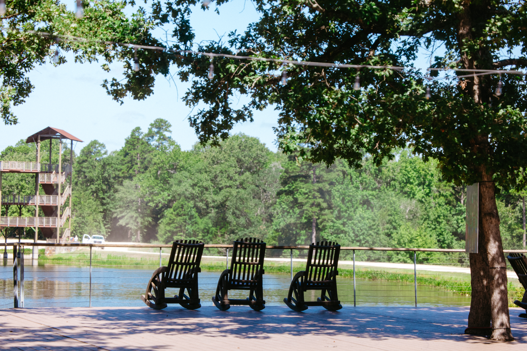 Three wooden rocking chairs on a lakeside deck shaded by tree branches with a wooden tower structure and forest in the background.
