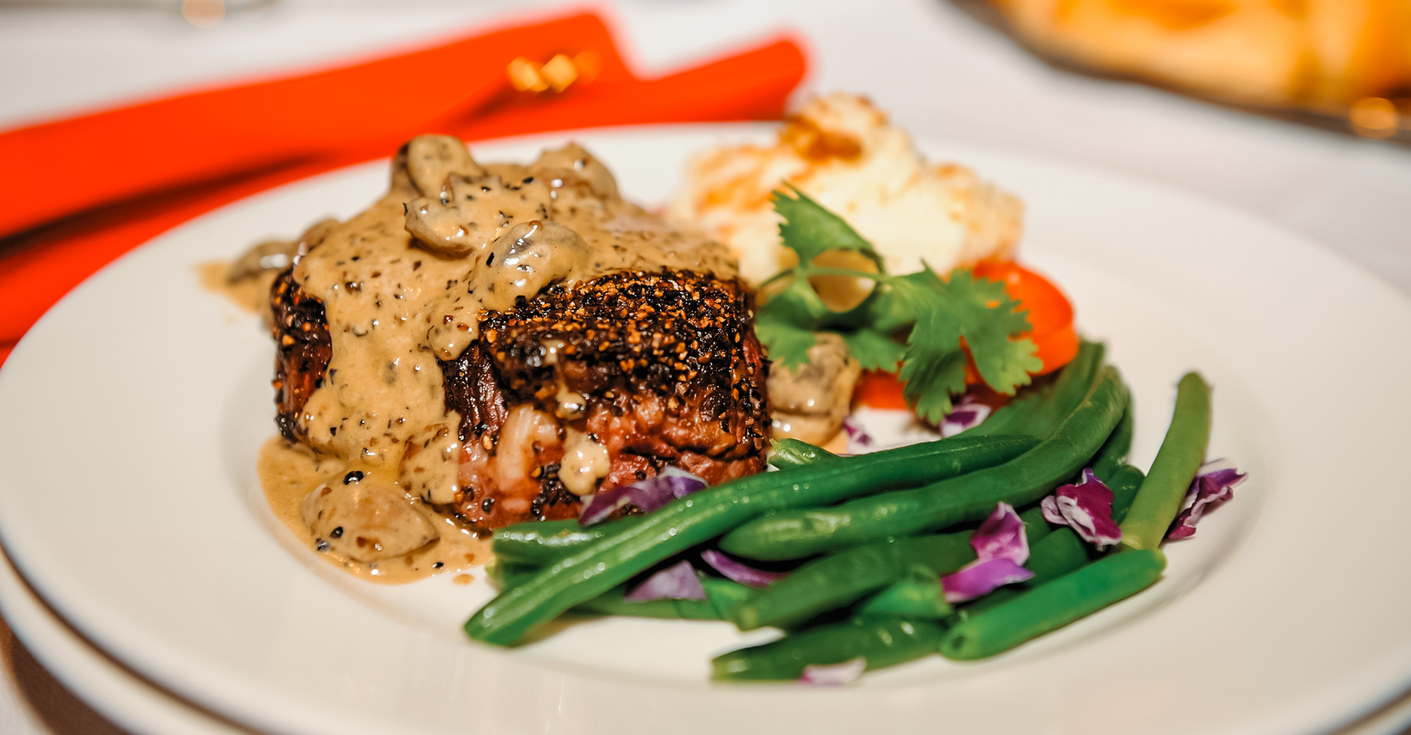 Grilled steak with creamy mushroom sauce, green beans, mashed potatoes, and garnished with cilantro and carrot slices on a white plate.