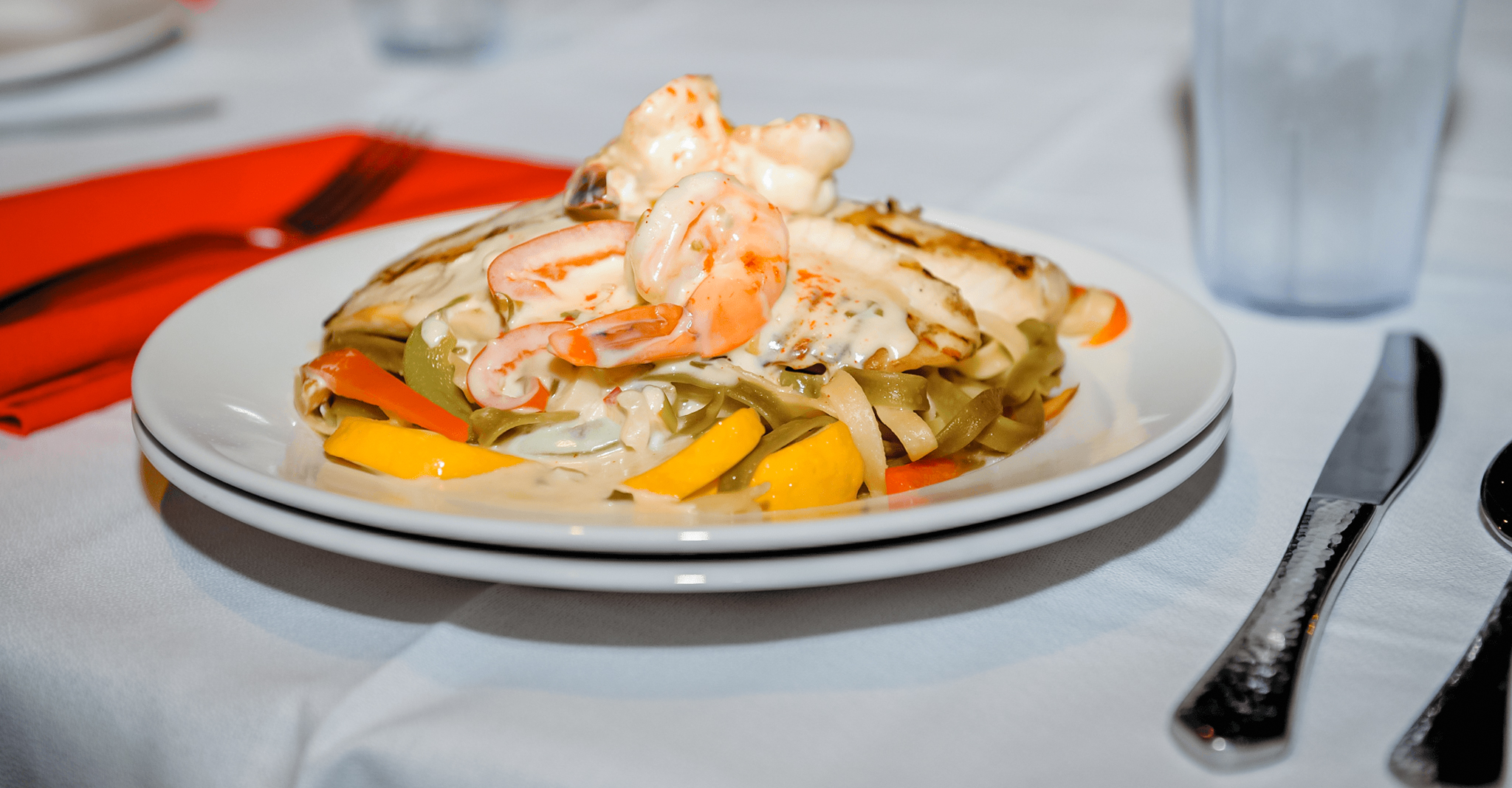 Plate of pasta with grilled chicken, shrimp, and vegetables in creamy sauce on a white tablecloth with cutlery and red napkin.
