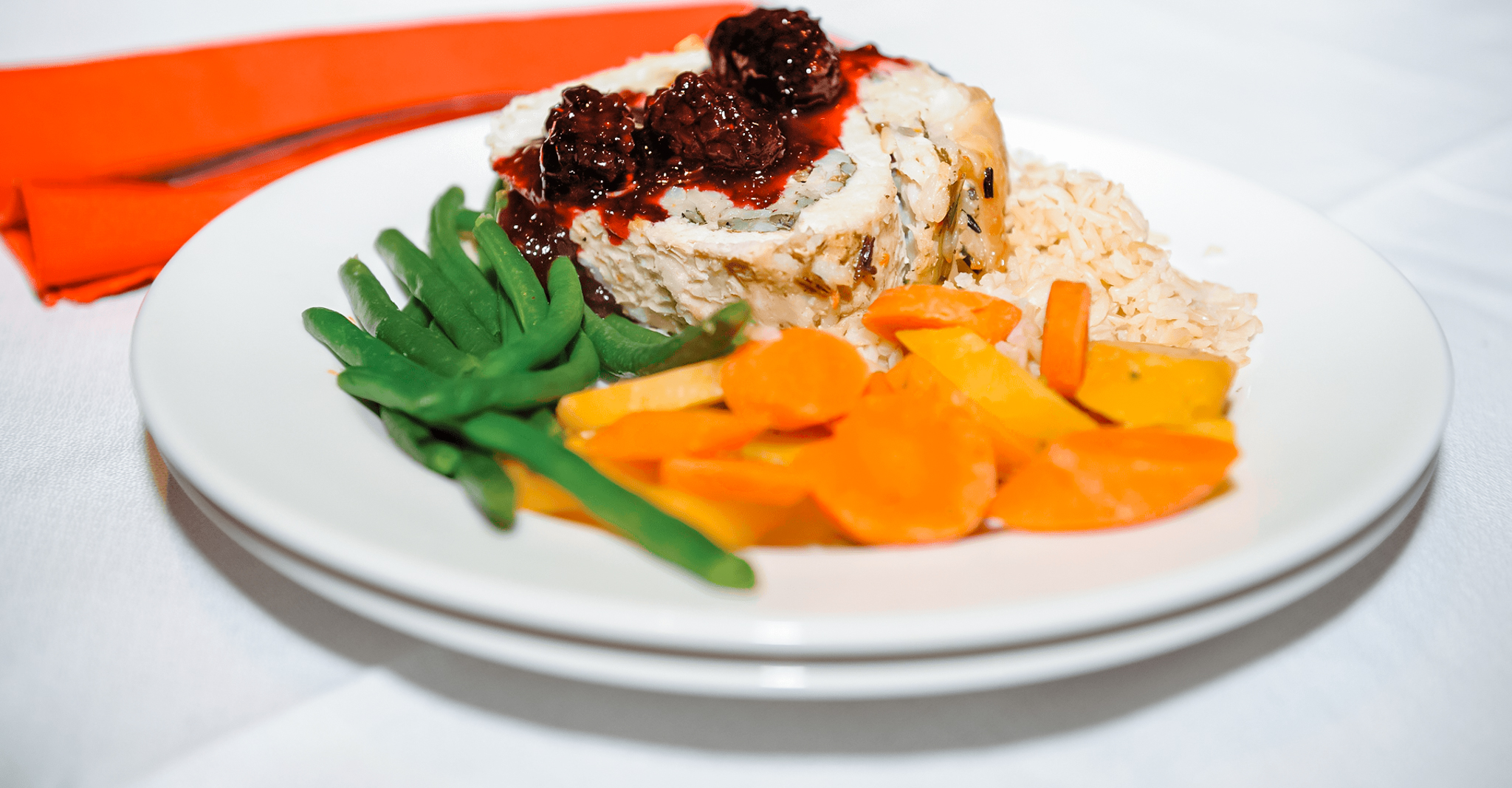 Plate with sliced turkey roll topped with cranberry sauce, green beans, cooked carrots, and wild rice on a white tablecloth.