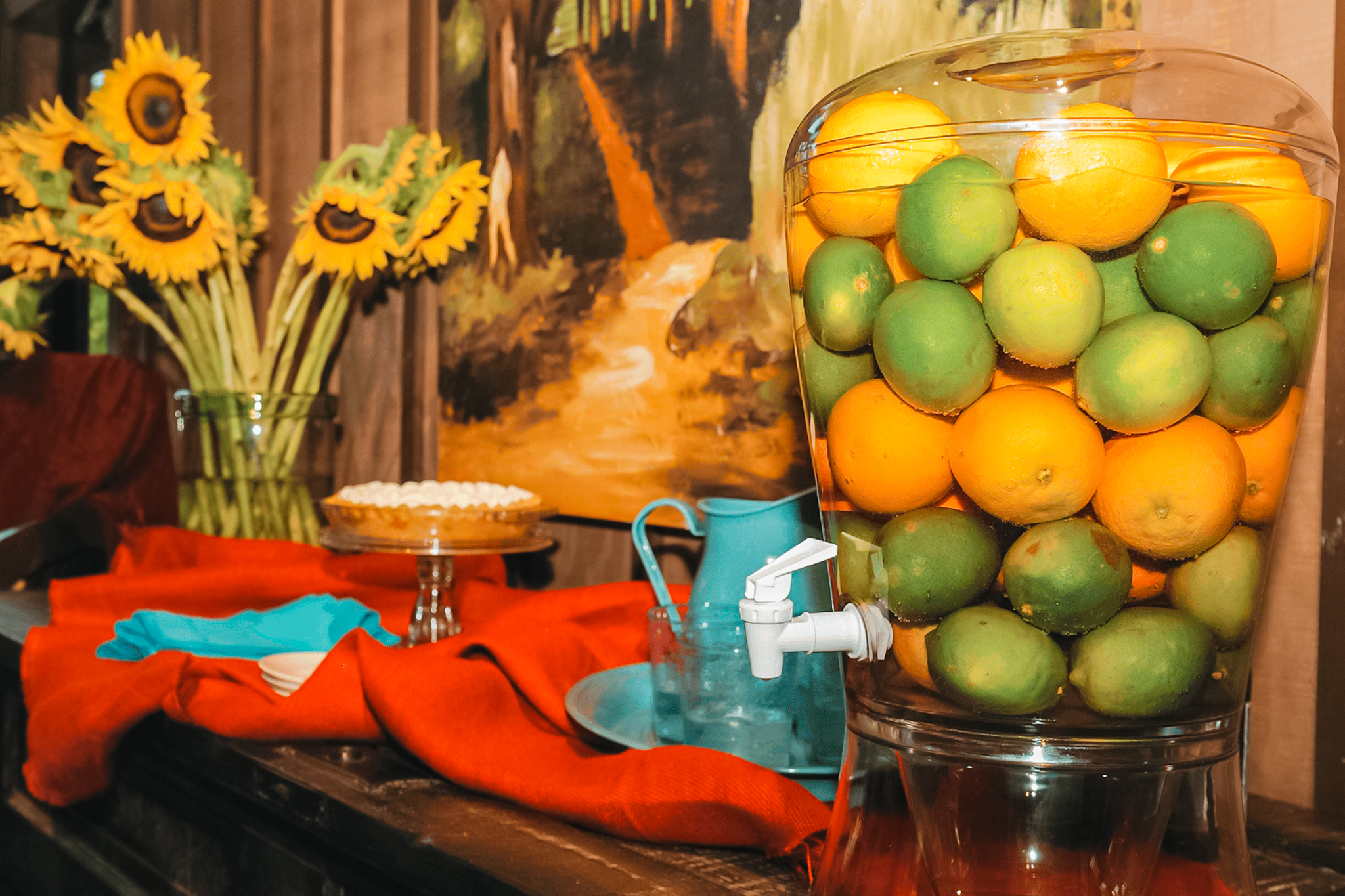 Glass beverage dispenser filled with limes and lemons on a table next to sunflowers, a pie, and drinkware.