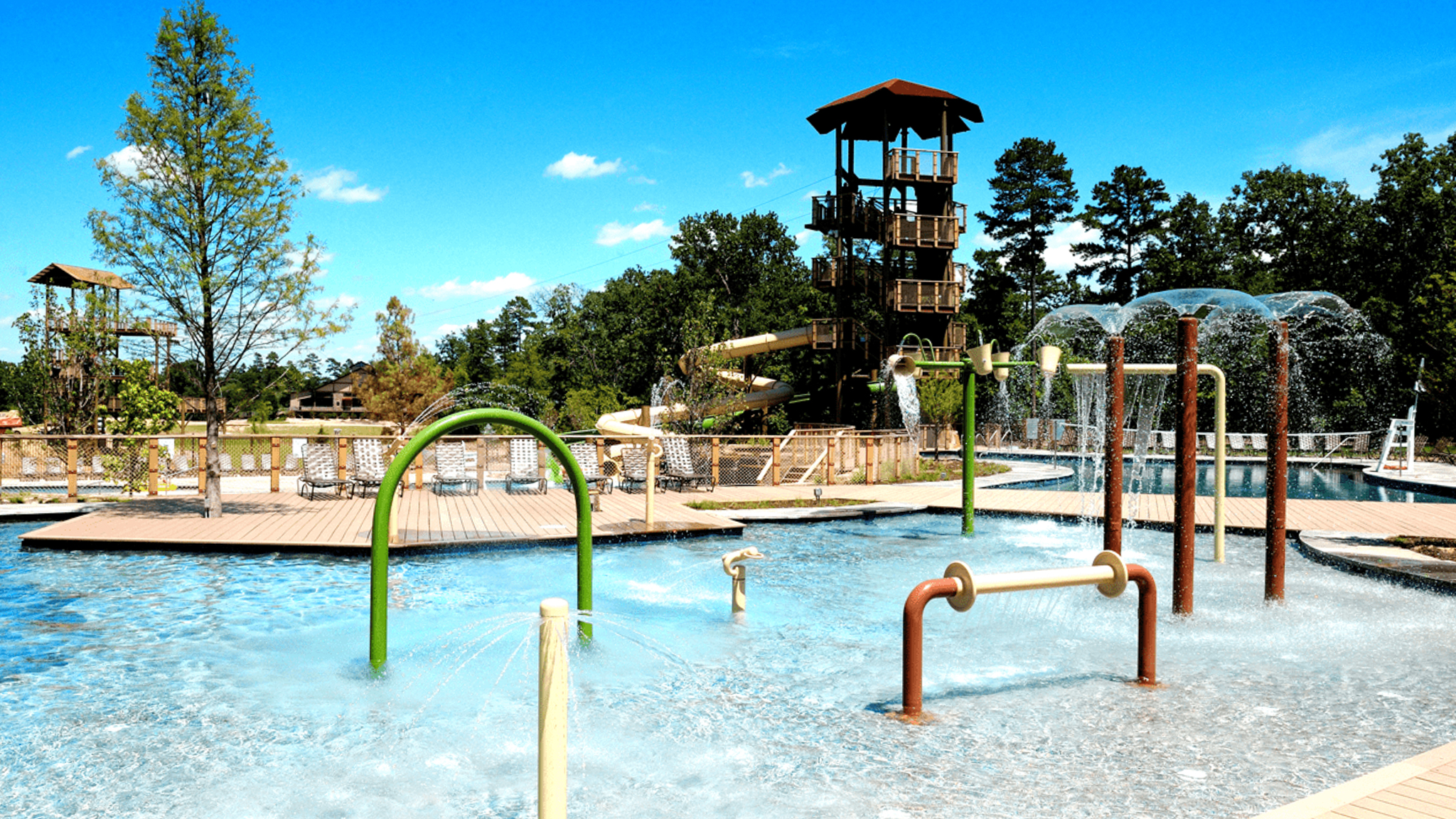 Outdoor water park with splash features, lounge chairs, and a tall wooden slide tower surrounded by trees.