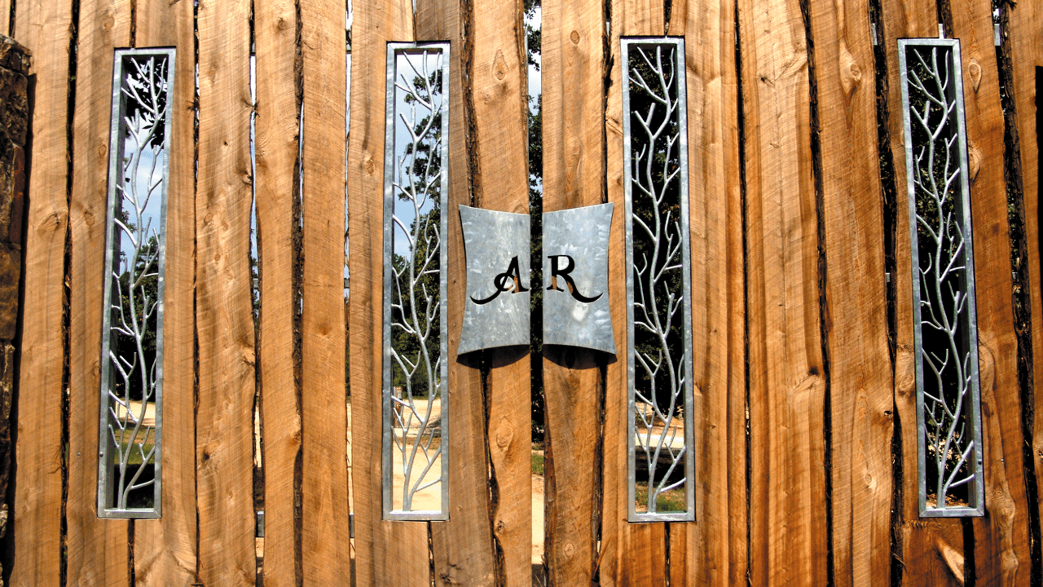 Wooden gate with vertical planks featuring narrow vertical windows with metal branch designs and a central metal plate displaying the letters 'A' and 'R'.
