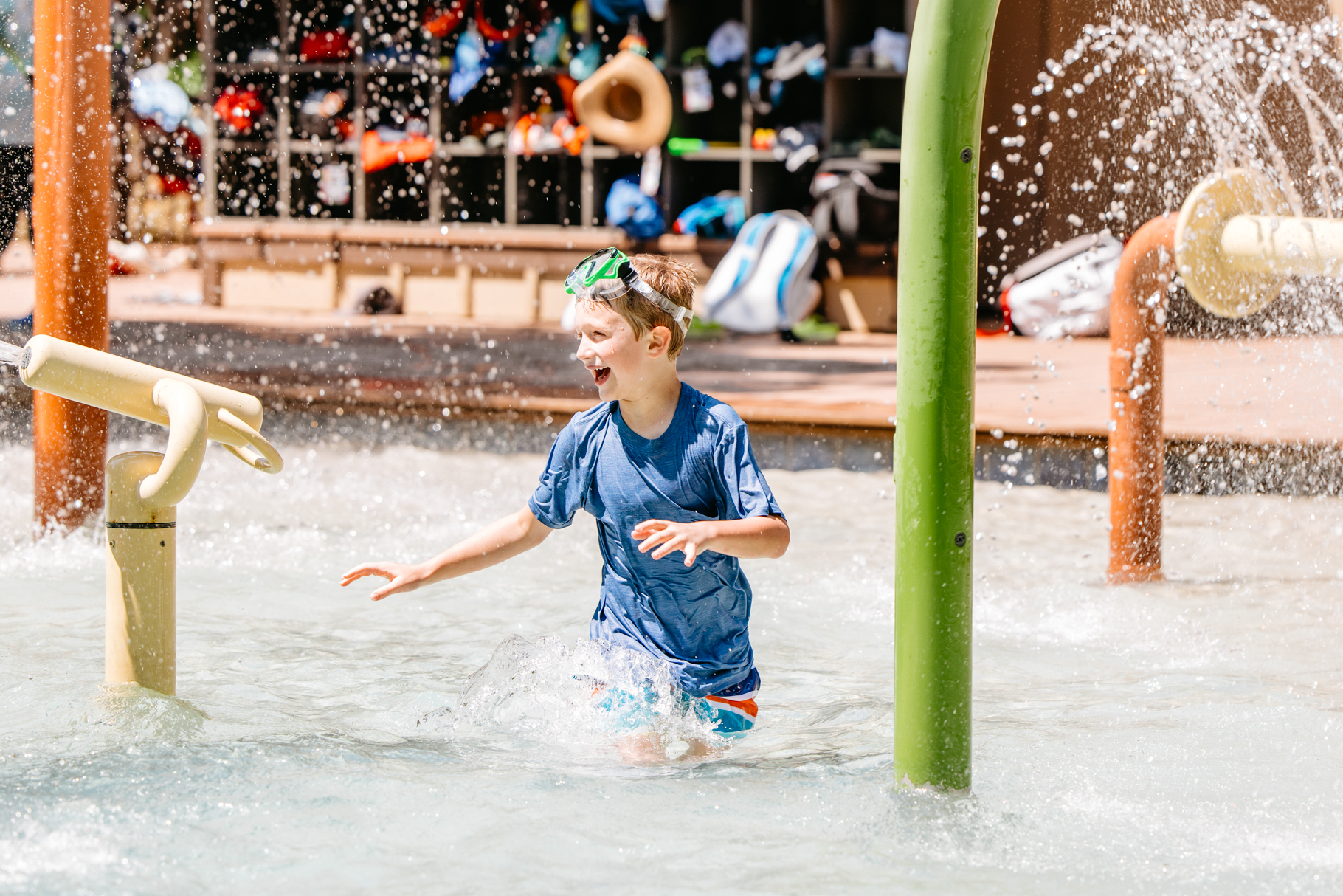 Young boy wearing a blue shirt and green goggles playing and splashing in a water park splash pad.