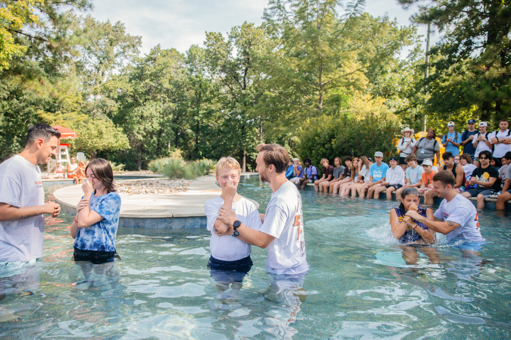 Two smiling men standing chest-deep in a pool during a baptism ceremony, with people seated by the poolside in the background.