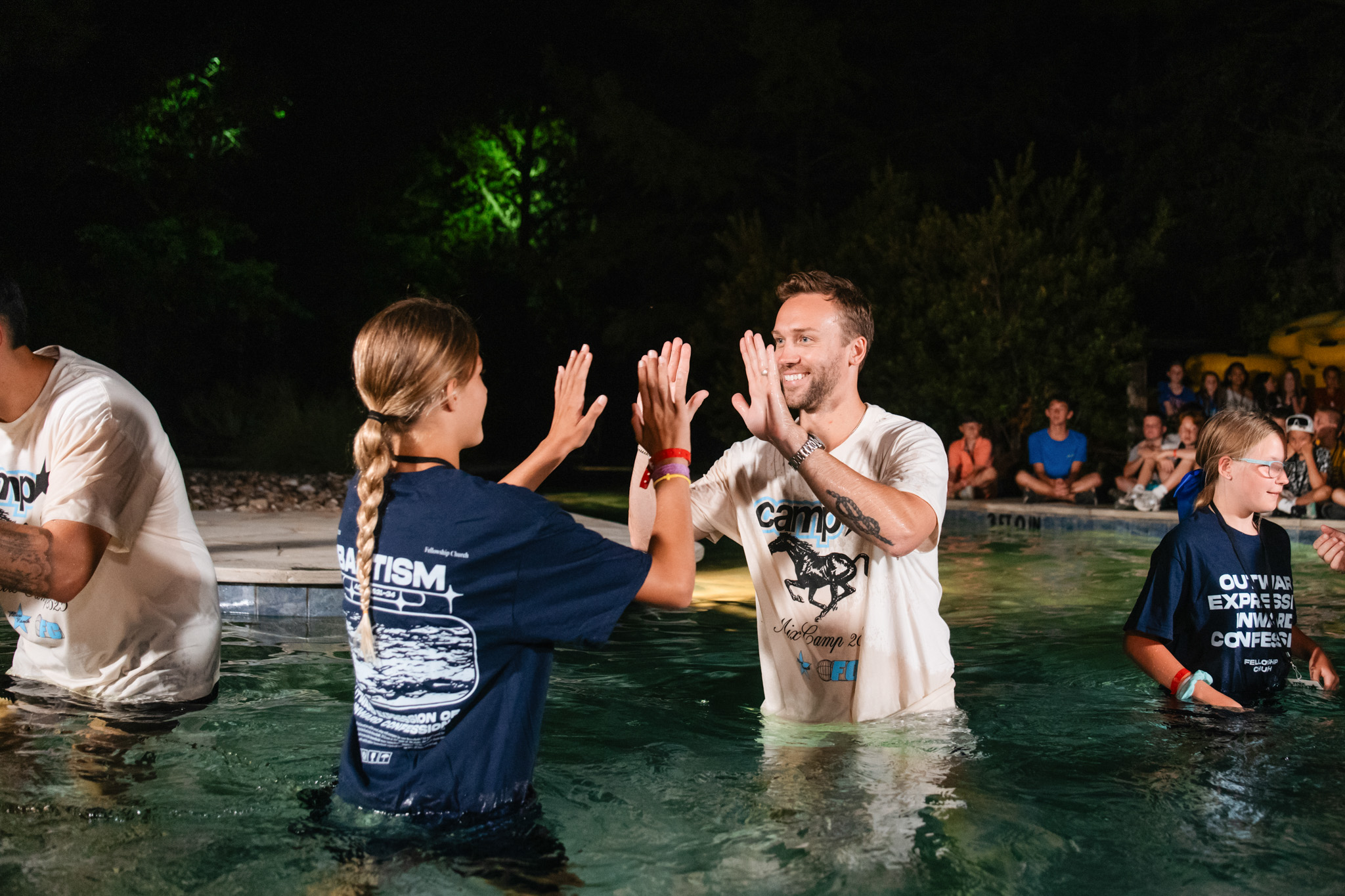 Two people standing waist-deep in a pool smiling, with a group of people sitting and standing around the poolside in casual summer clothing.
