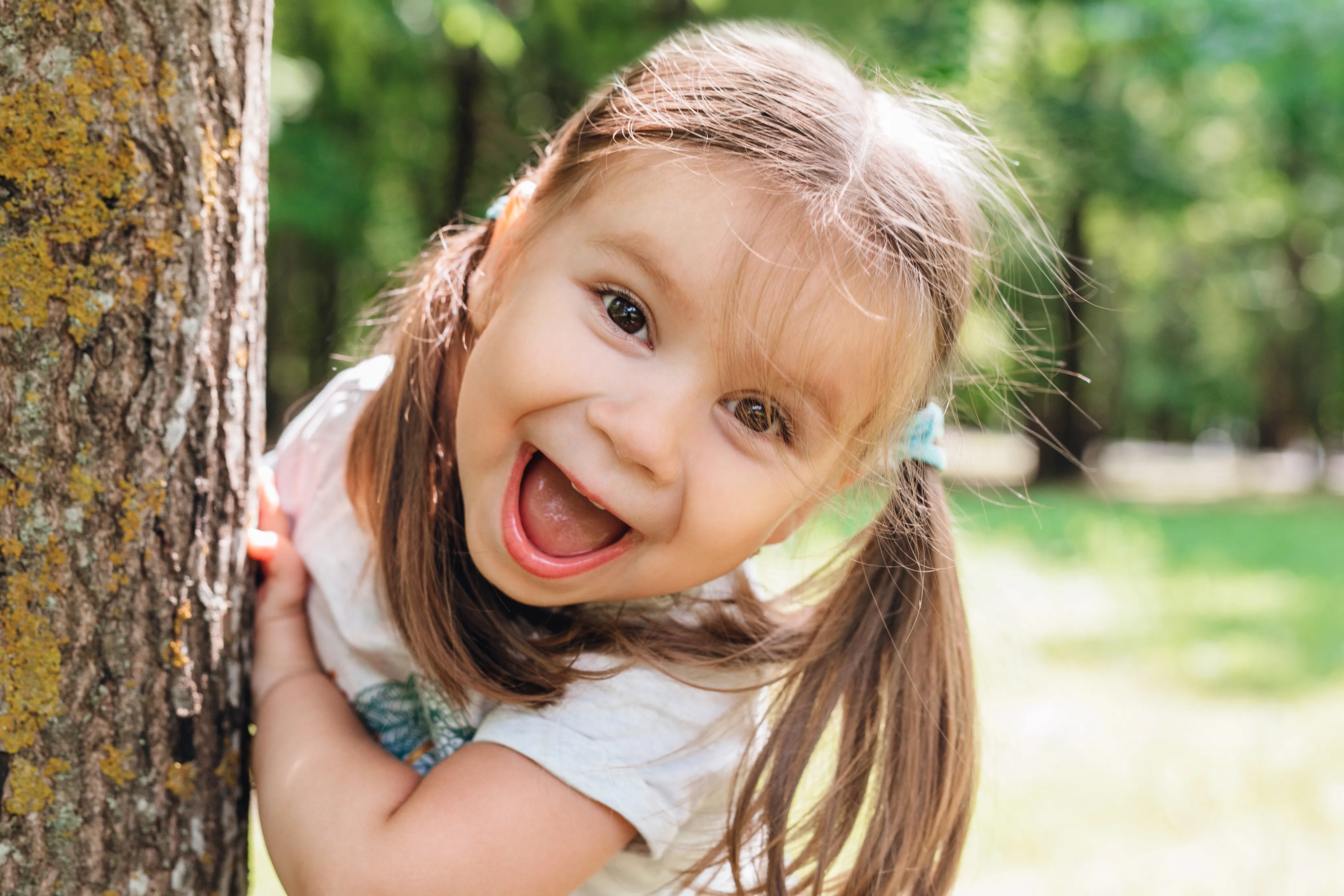 A little girl is peeking from behind a tree and smiling. 