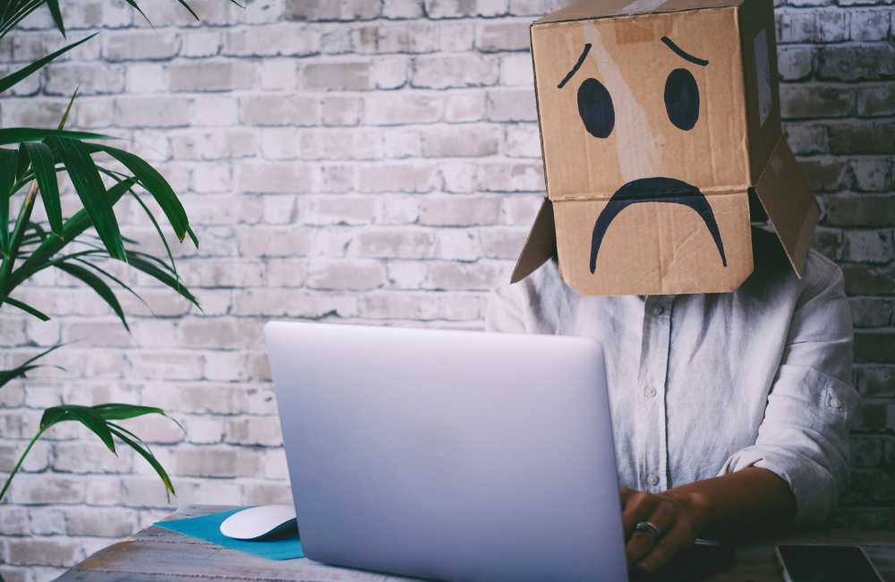 A person at a computer wearing a cardboard box on their head with a sad face on it
