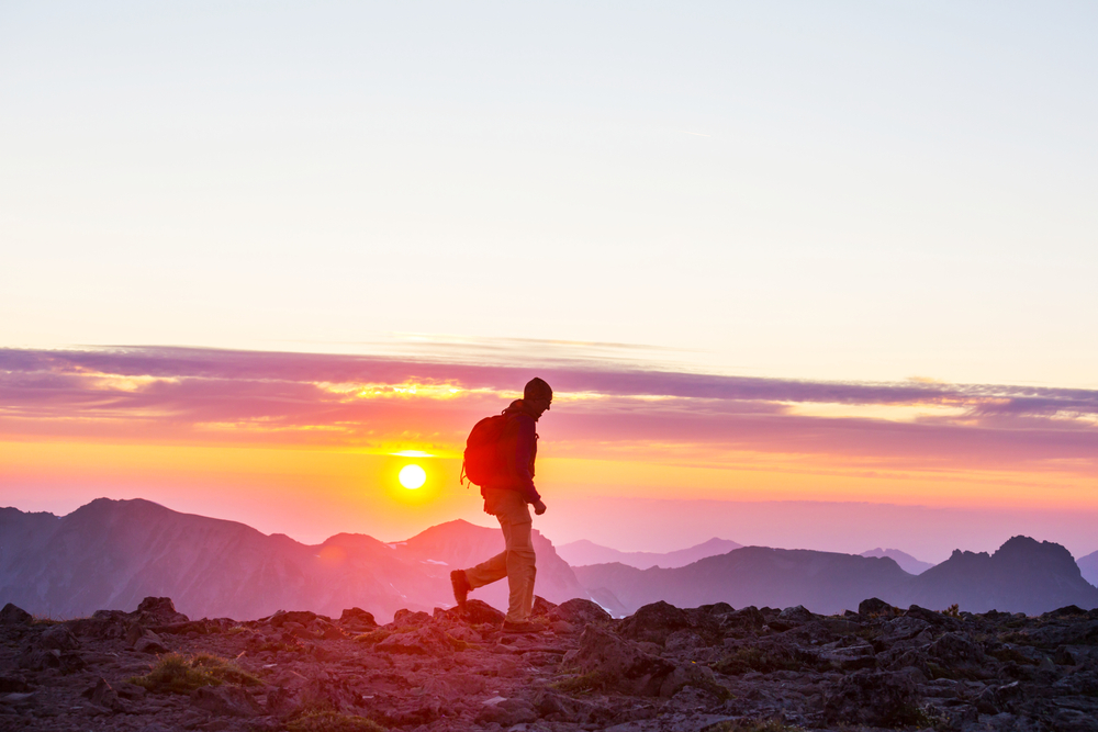 A hiker walking along a mountain top with a sunrise in the background.