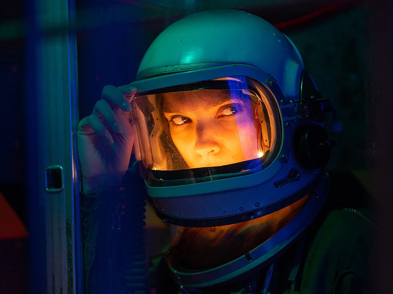 A futuristic woman wearing a retro astronaut helmet. Inside the helmet has a warm glow while the surrounding is tinted a cold blue.