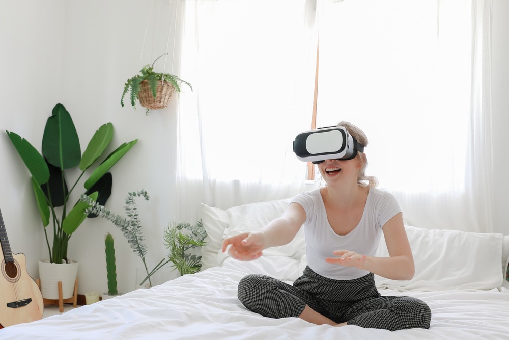 A woman sitting on a bed using a virtual reality headset, reaching out to something unseen.