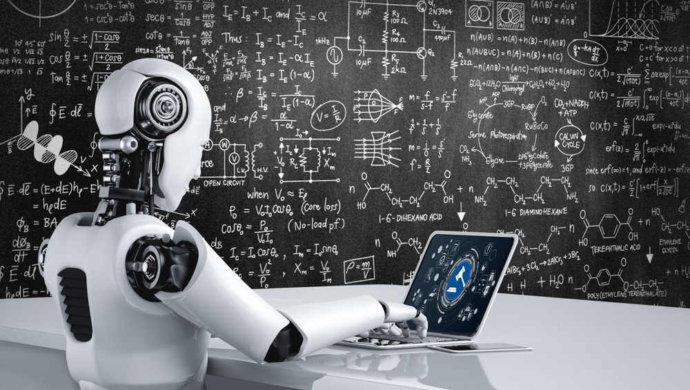 An AI robot sits at a laptop with a blackboard covered in mathematics behind it.