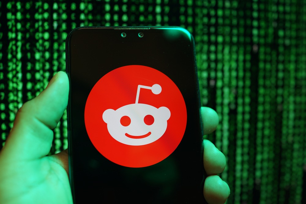 A hand holding a phone displaying the Reddit logo. There is computer code in the background.