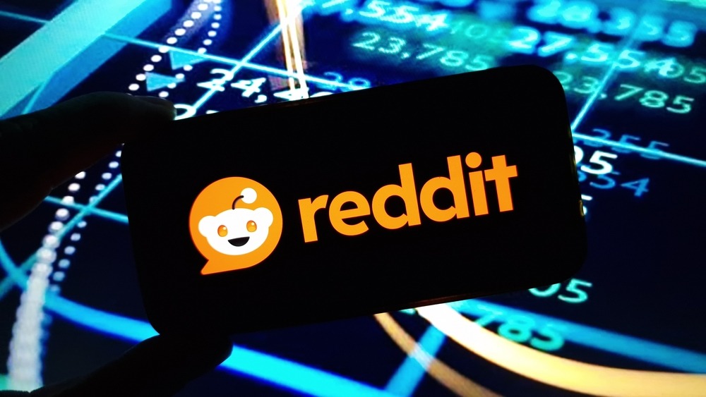The Reddit logo displayed on a phone with computer analytics and code in the background. 