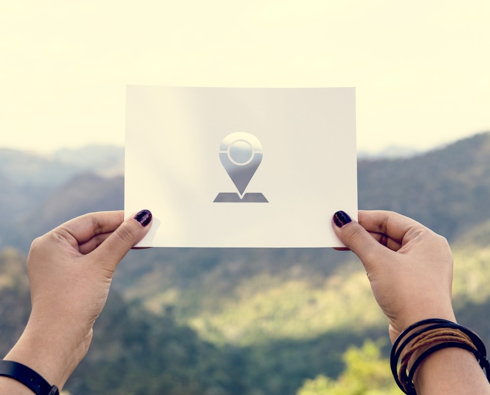 A location pin cut out of paper and held by hands in front of a natural landscape.