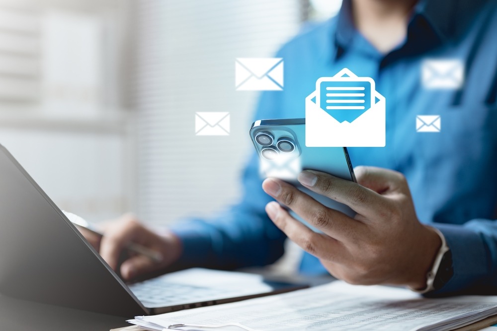 A person working at a desk on a laptop out of focus holding their phone that is in focus. There are email graphics hovering around the phone with one indicating an opened email.