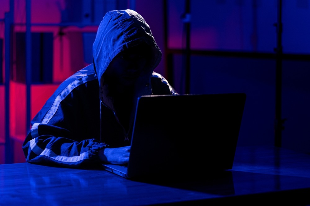 A person at laptop with their hood up with their face darkened by shadow to appear faceless.