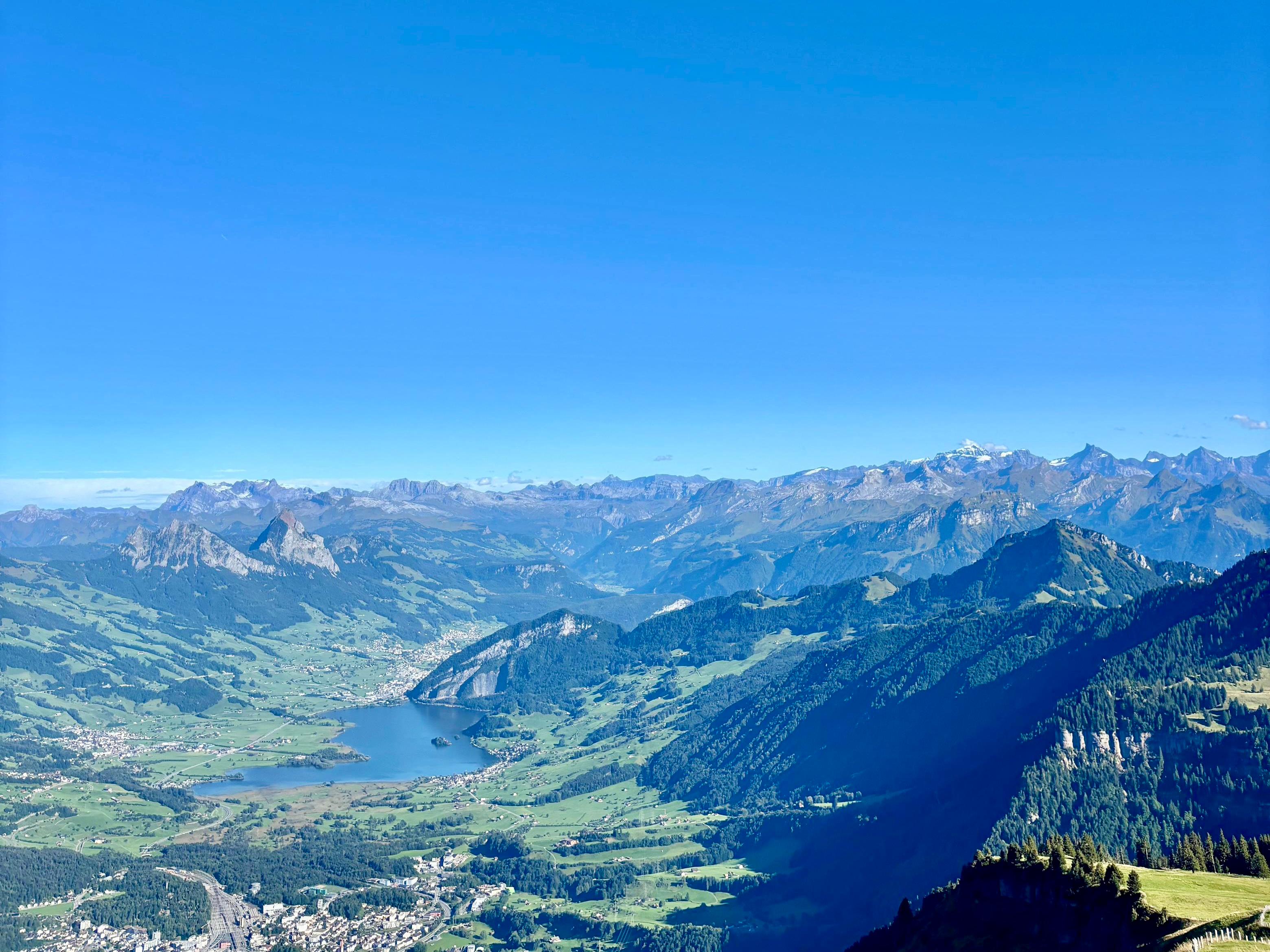 Ausblick auf die Alpenkette, einem See und grünbewachsenen Hängen bei strahlend blauen Himmel.